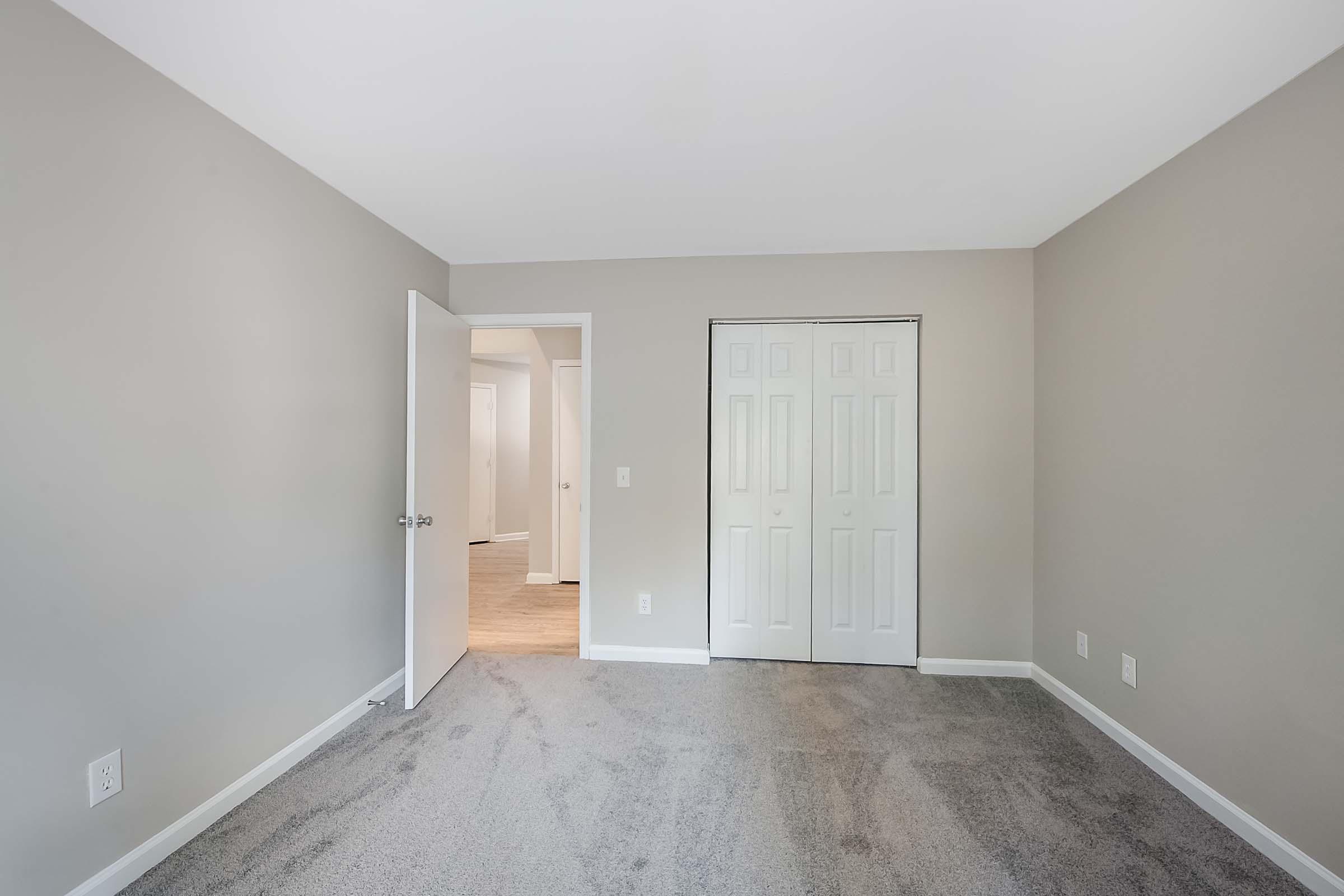 A clean, empty room with light gray walls and carpeted flooring. There is a door leading to another room, and a closet with double doors on one side. Natural light from an adjacent room brightens the space, creating a minimalist and inviting atmosphere.