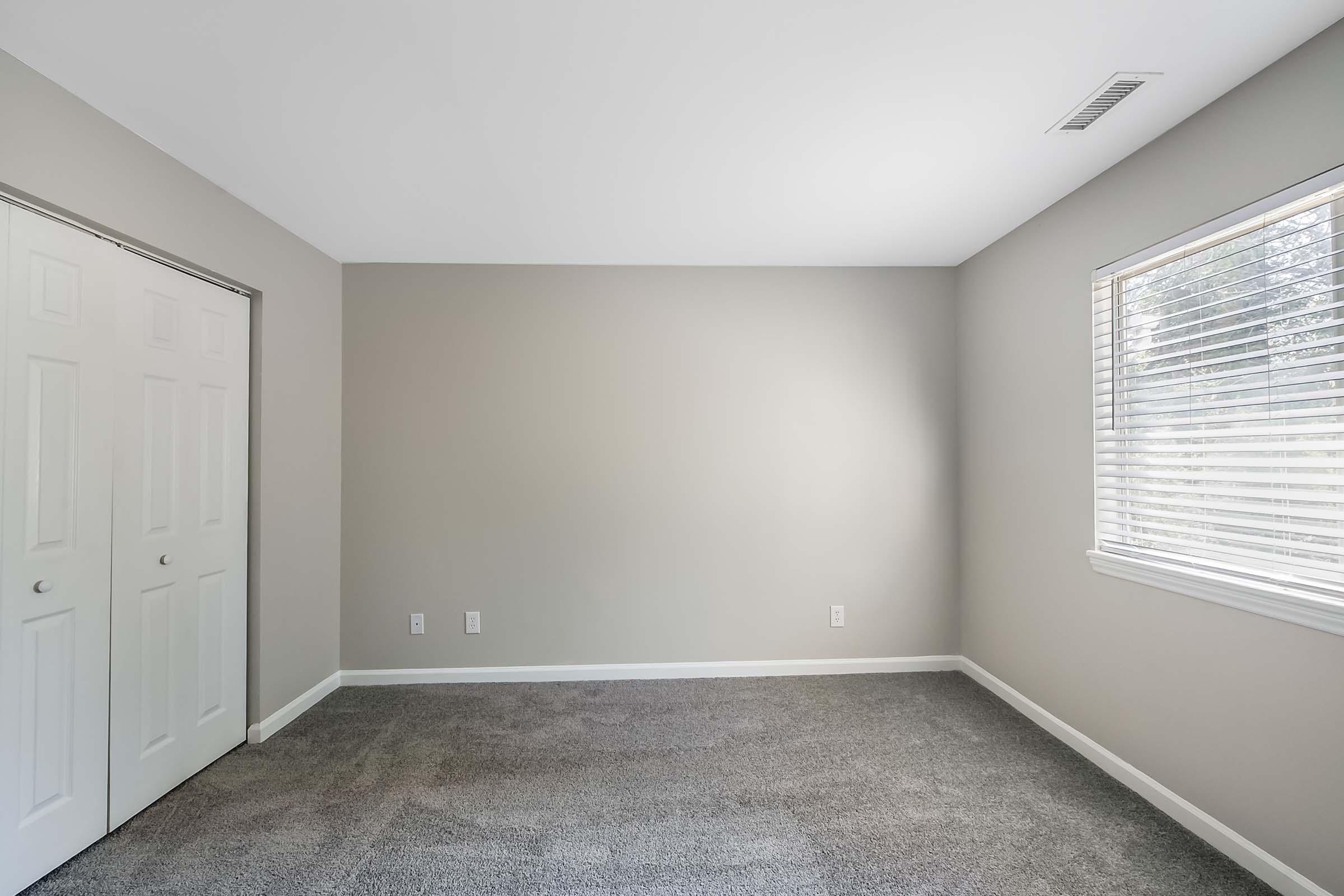 Empty room with light gray walls and carpeted floor. There is a window with blinds on the right, letting in natural light, and a double closet with white sliding doors on the left. The room is unfurnished, creating a clean and spacious atmosphere.