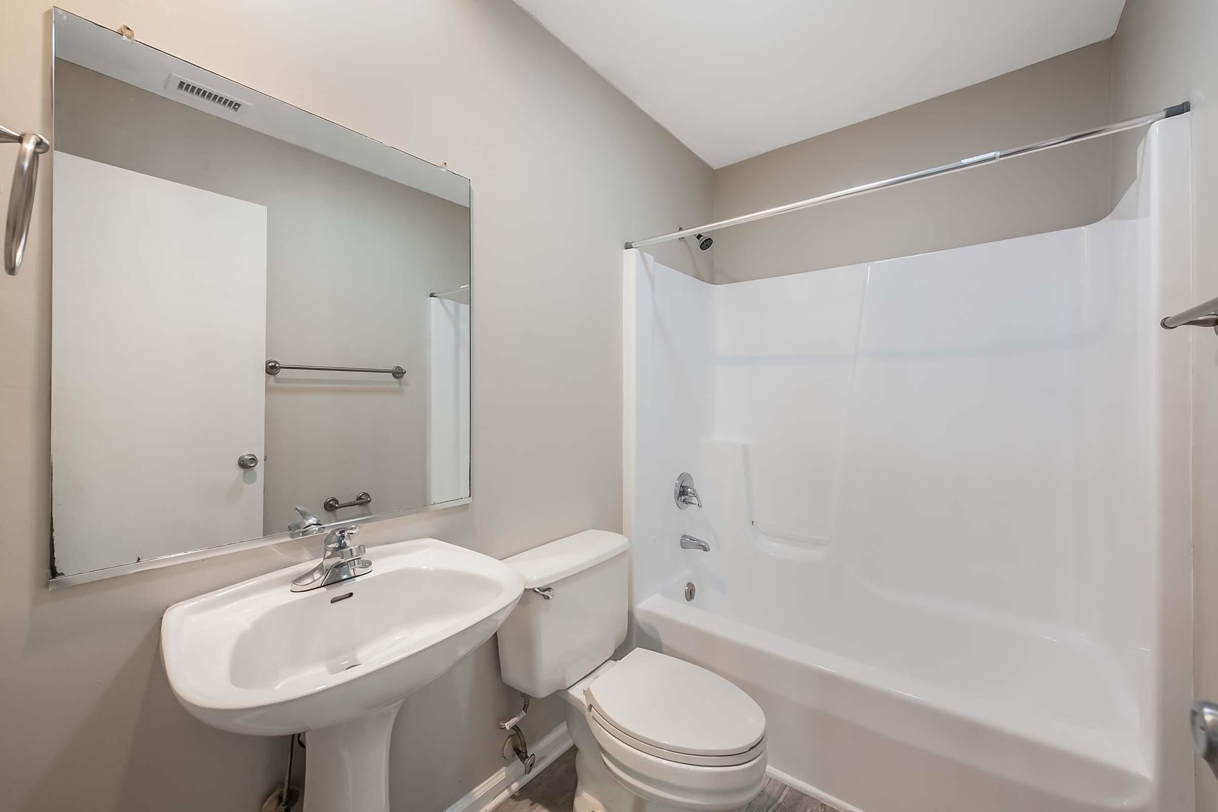 A clean and modern bathroom featuring a white bathtub with a shower, a white sink, and a toilet. The walls are painted a neutral gray, and there is a large mirror above the sink. A towel rack is mounted on the wall, and the overall space appears well-maintained and bright.