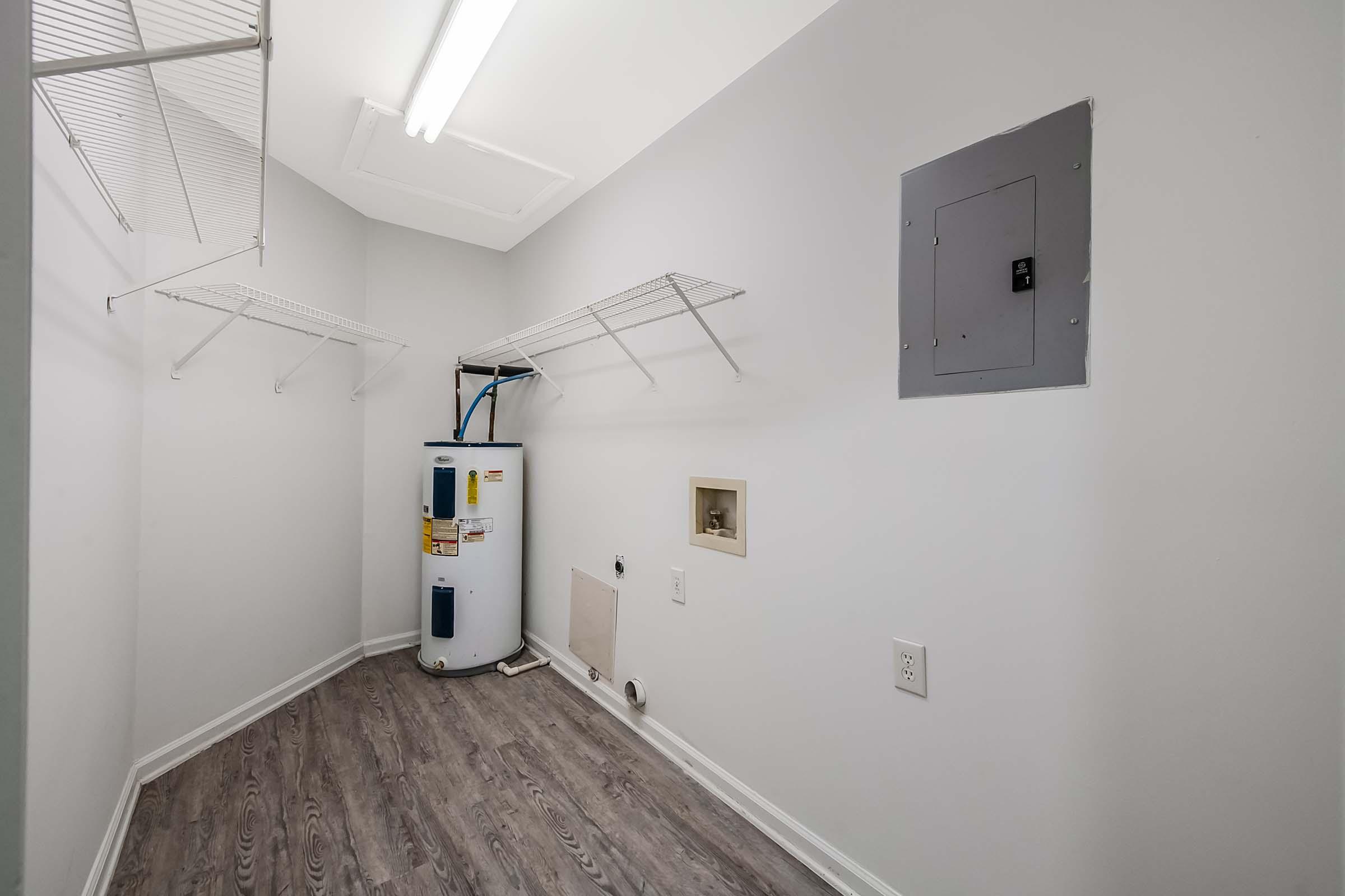 A clean, empty laundry room featuring a water heater on the left side. The walls are painted light gray, and there are several white wire shelves mounted on the walls. The floor has a wood-like laminate finish, and there is a gray electrical panel on the right side. Overall, the space is bright and spacious.