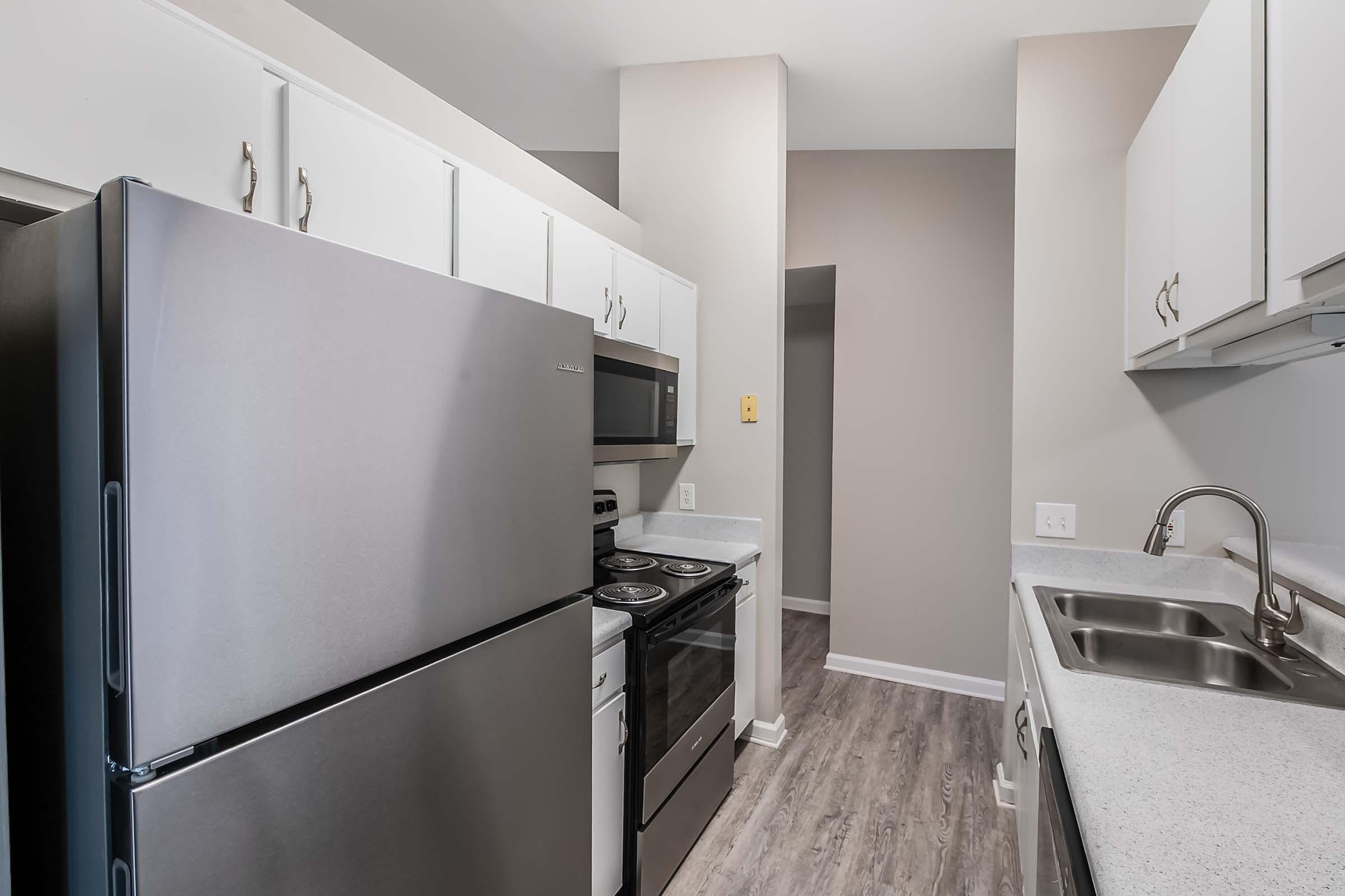 A modern kitchen featuring stainless steel appliances including a refrigerator and an oven, with white cabinetry and a gray countertop. The space has light-colored walls and wood-style flooring, creating a clean and contemporary look. A sink is installed next to the countertop for functionality.