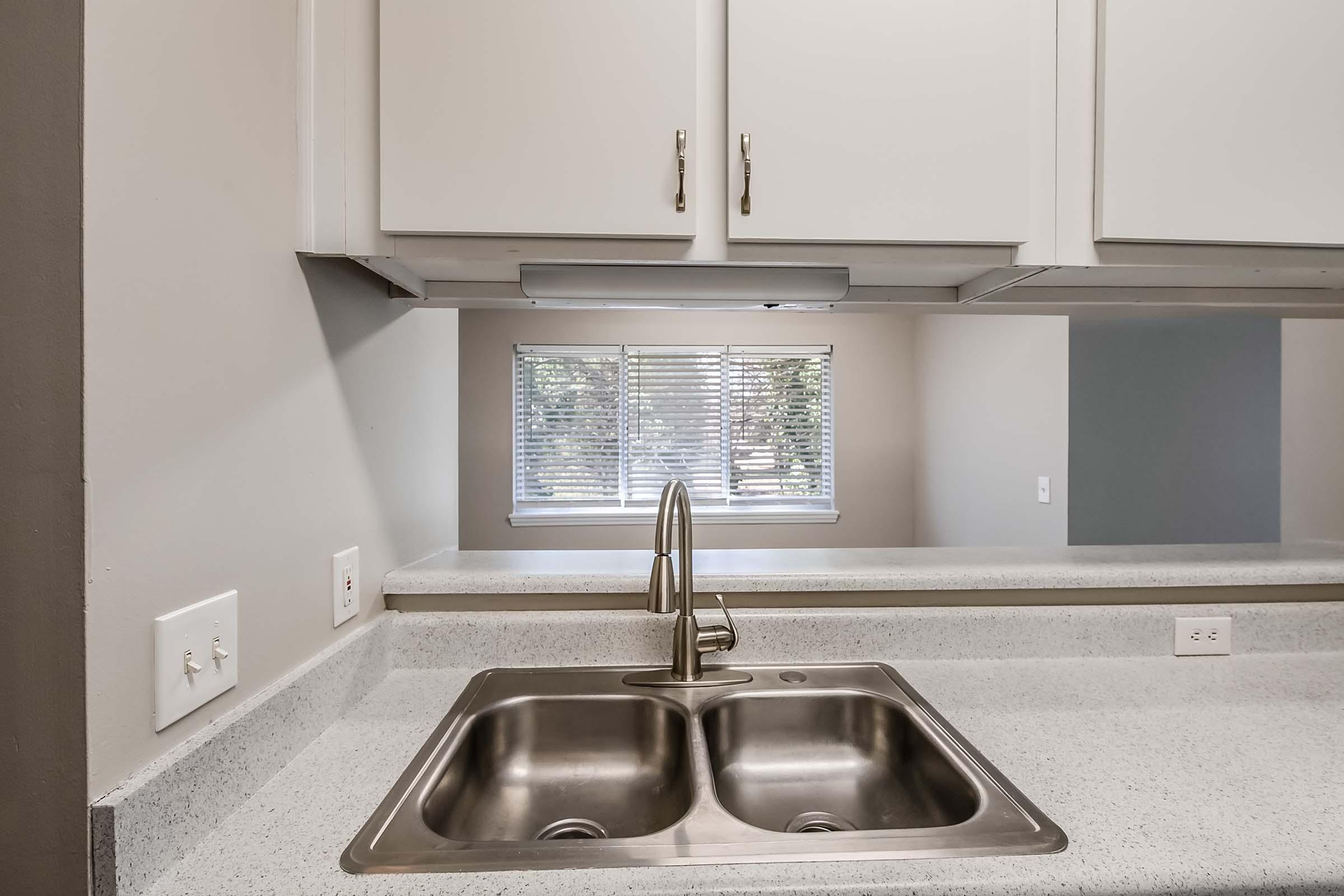 A modern kitchen with a stainless steel double sink and a light gray countertop. White cabinetry above the sink features sleek handles. In the background, a window with horizontal blinds allows natural light to enter the space, creating a bright and airy atmosphere.