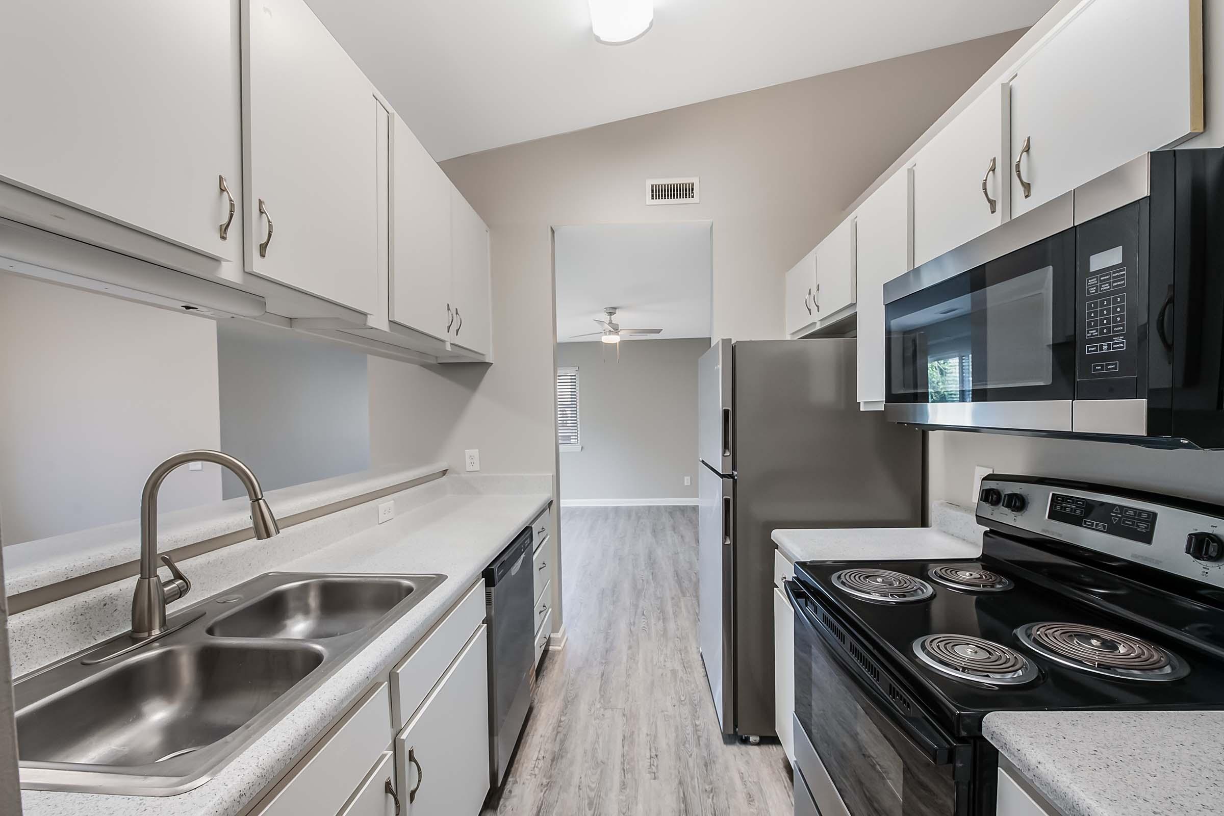 Modern kitchen featuring white cabinetry, a stainless steel sink, and a double basin. Equipped with a microwave, a black electric stove, and a side-by-side refrigerator. The flooring is light-colored, and there are windows allowing natural light to enter, creating an open and inviting space.