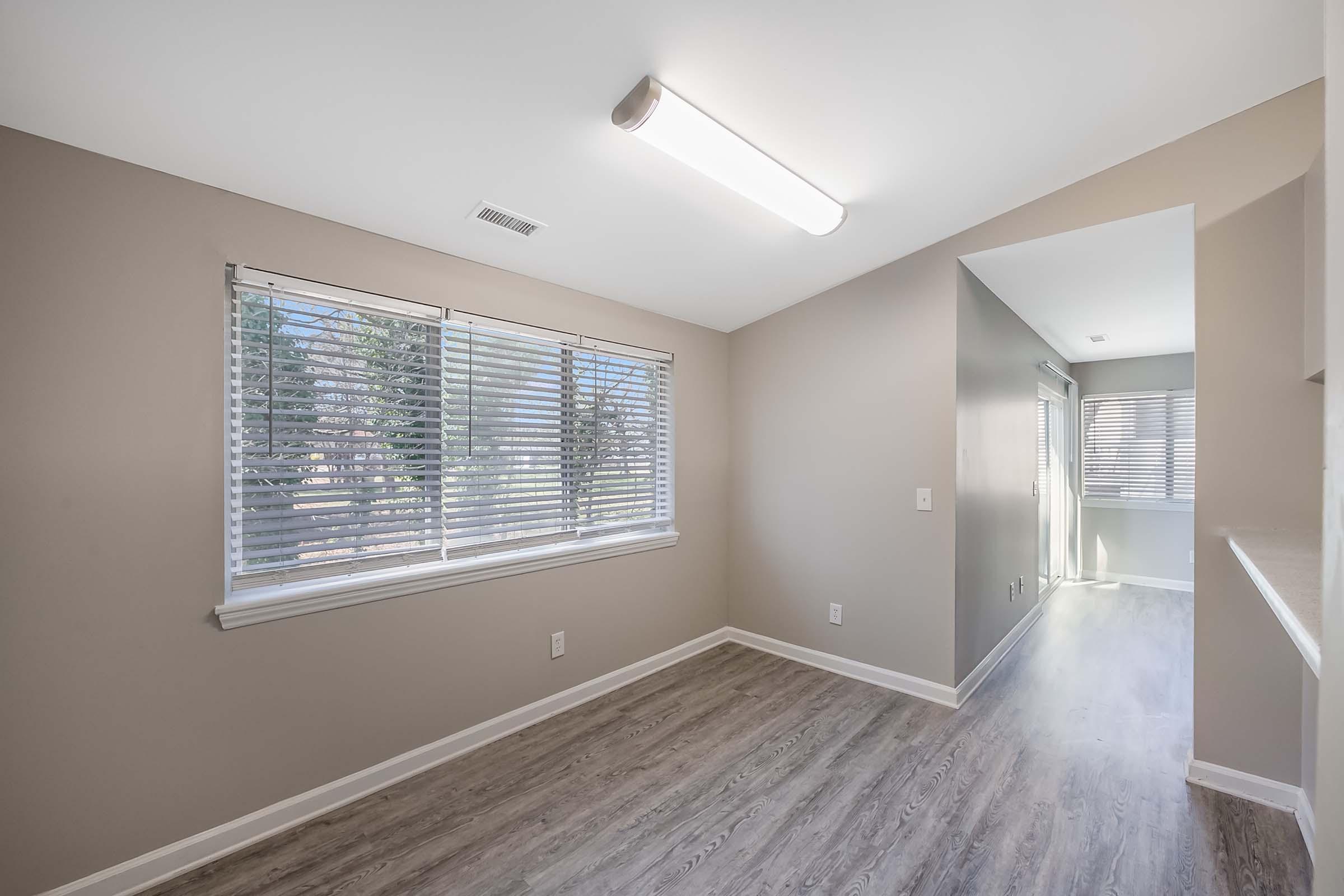 A well-lit corner room featuring two windows with blinds, light beige walls, and laminate flooring. The space appears open and airy, with a ceiling light fixture and a small adjacent area visible in the background, suggesting a welcoming environment suitable for various uses.