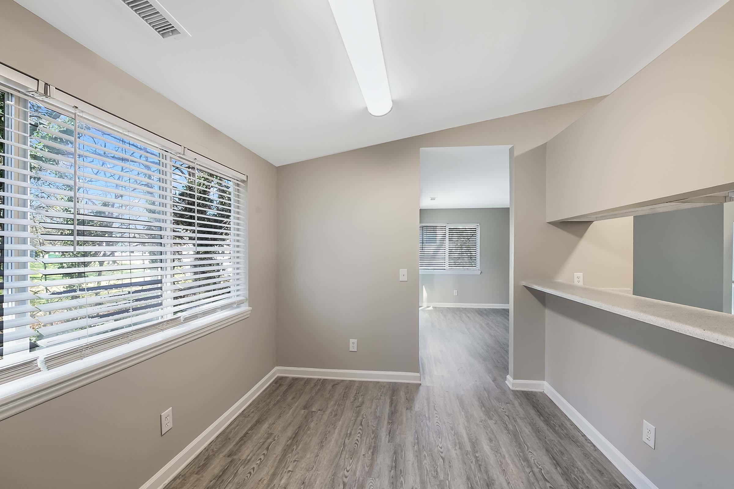 A well-lit, modern interior space featuring a light-colored wall, large windows with blinds, and a view of another room. The floor is covered in wood-like laminate, and there is a small counter that separates the kitchen area from the living space. Bright natural light fills the room.