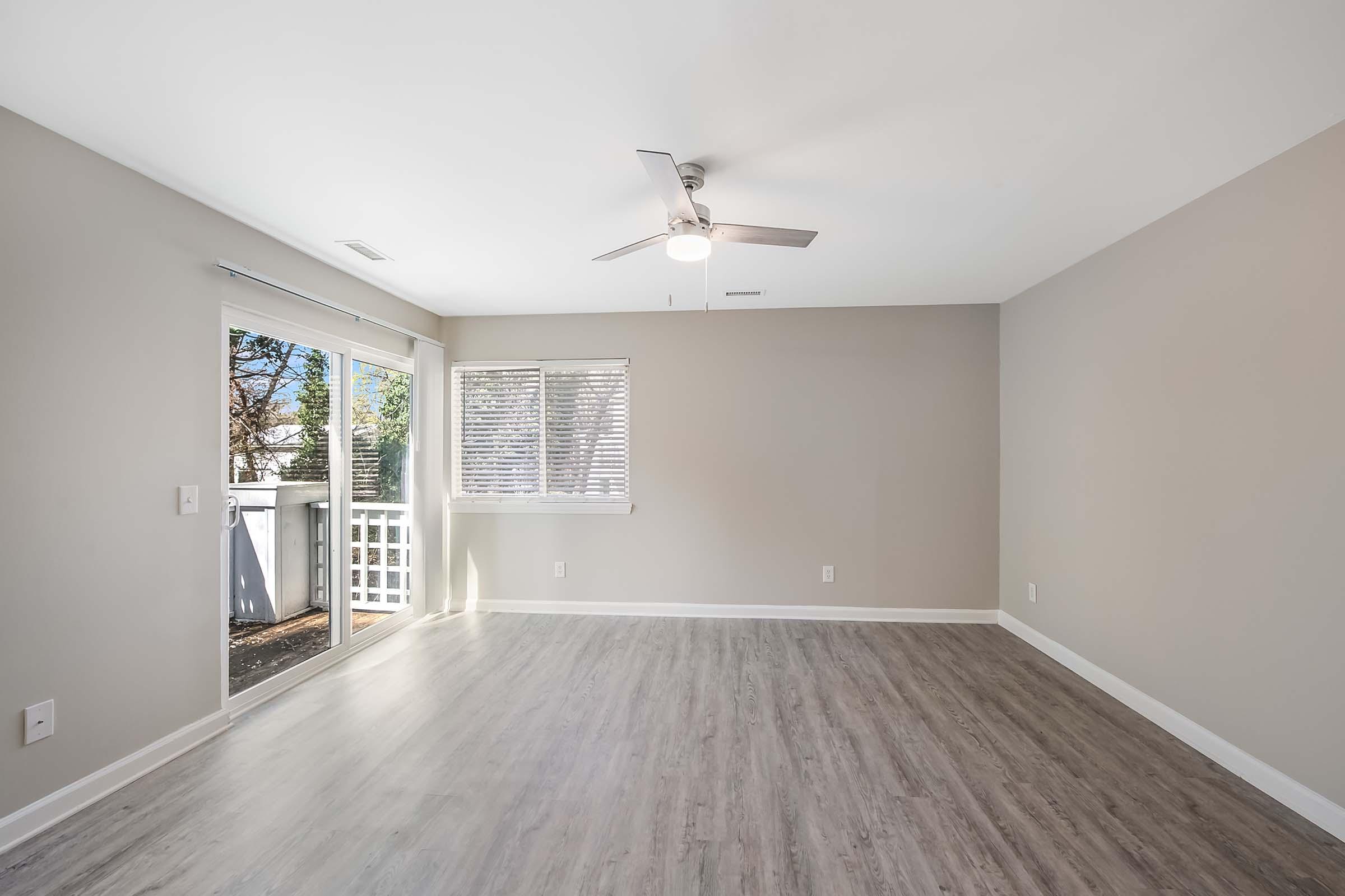 A spacious, empty room featuring light gray walls and a modern ceiling fan. Large windows allow natural light to fill the space, with a sliding glass door leading to an outdoor area. The floor is covered in light wood-like laminate, creating a bright and airy atmosphere.