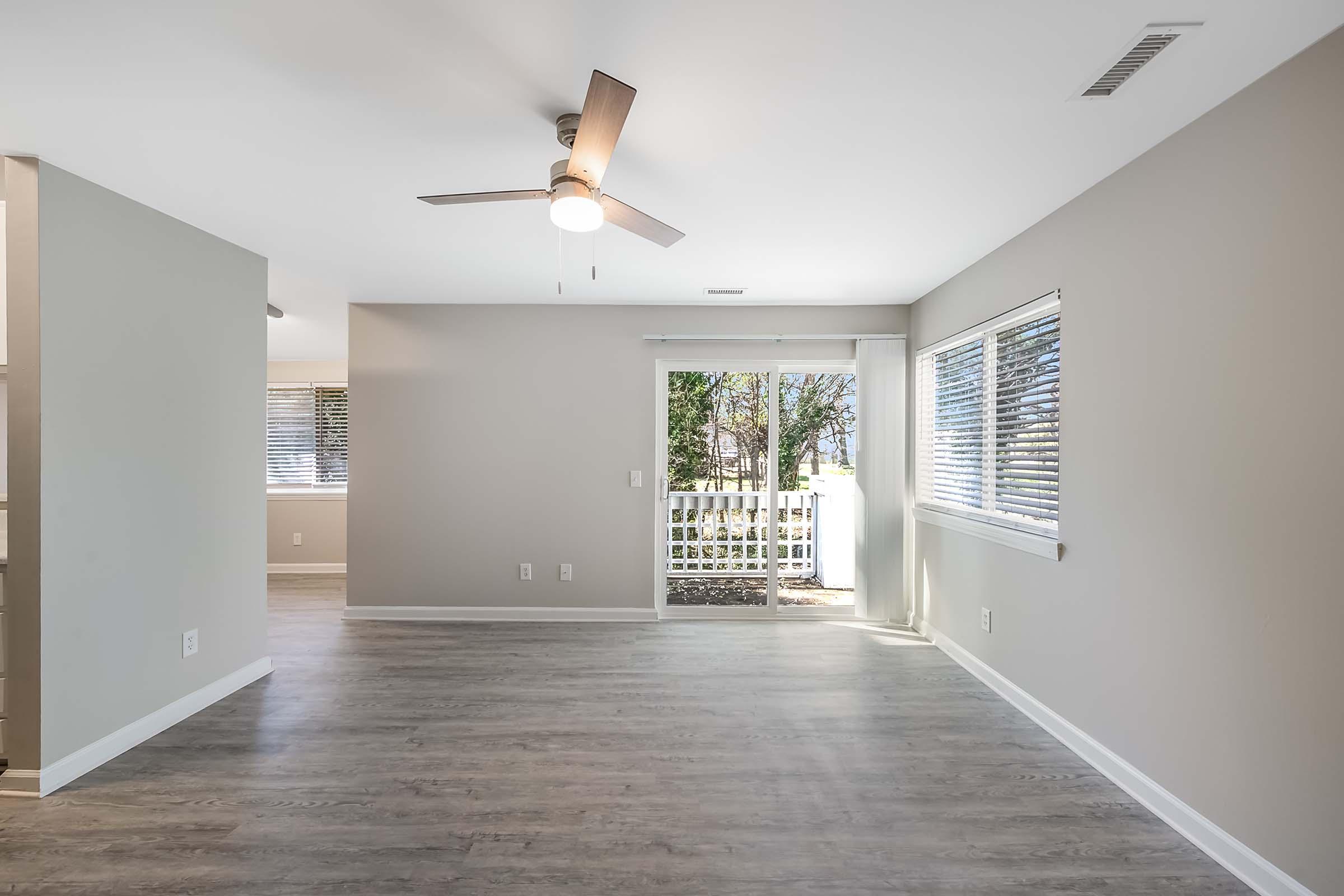 Spacious, light-filled living area with light gray walls and modern ceiling fan. Large windows with blinds offer a view of the outdoors. Wood-like flooring adds warmth to the space, which opens to a patio area through glass doors.