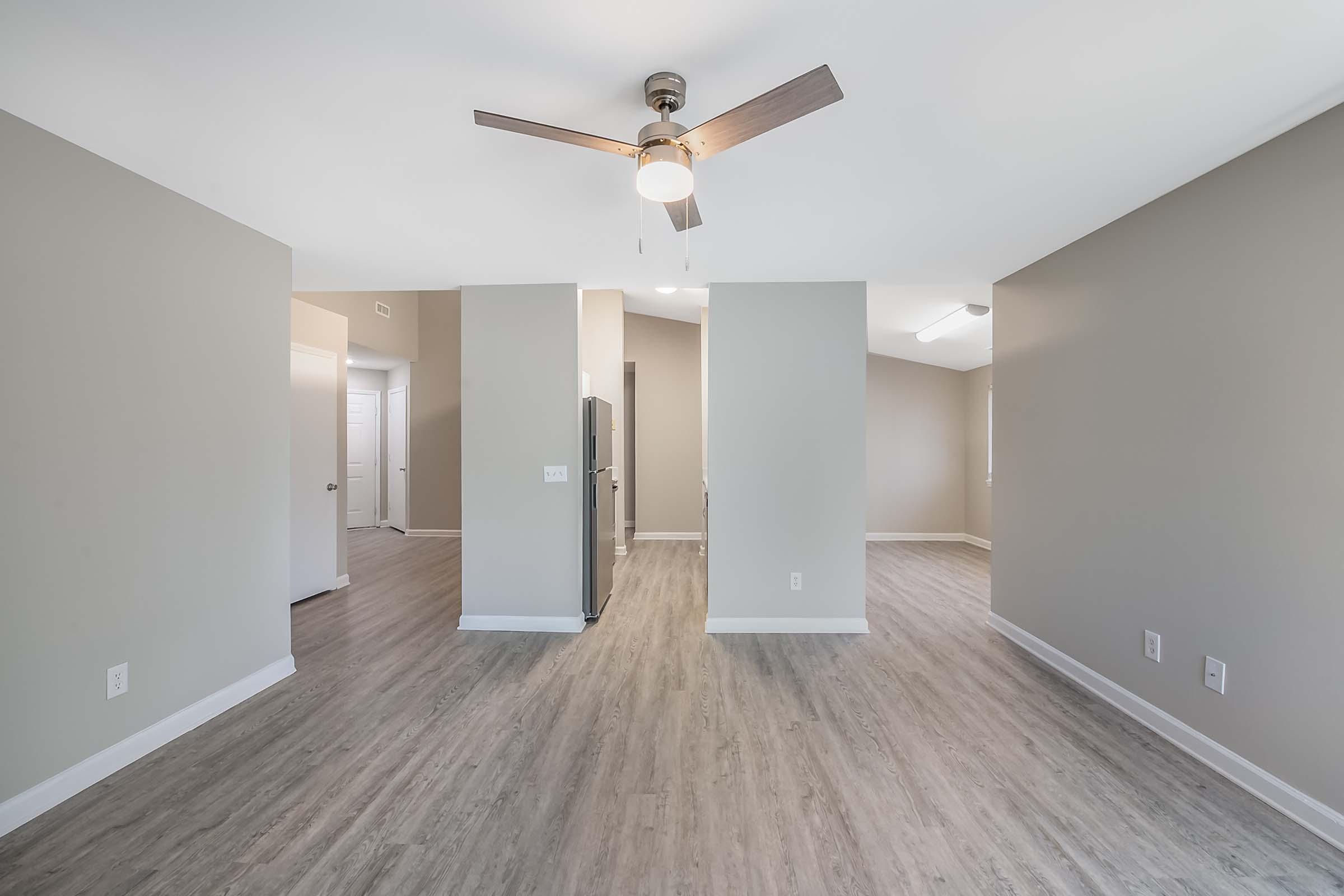 A spacious, well-lit living area featuring a modern ceiling fan, light-colored walls, and laminate flooring. The room opens to a kitchen area with a refrigerator visible. There are doorways leading to other rooms, creating a welcoming and airy atmosphere.