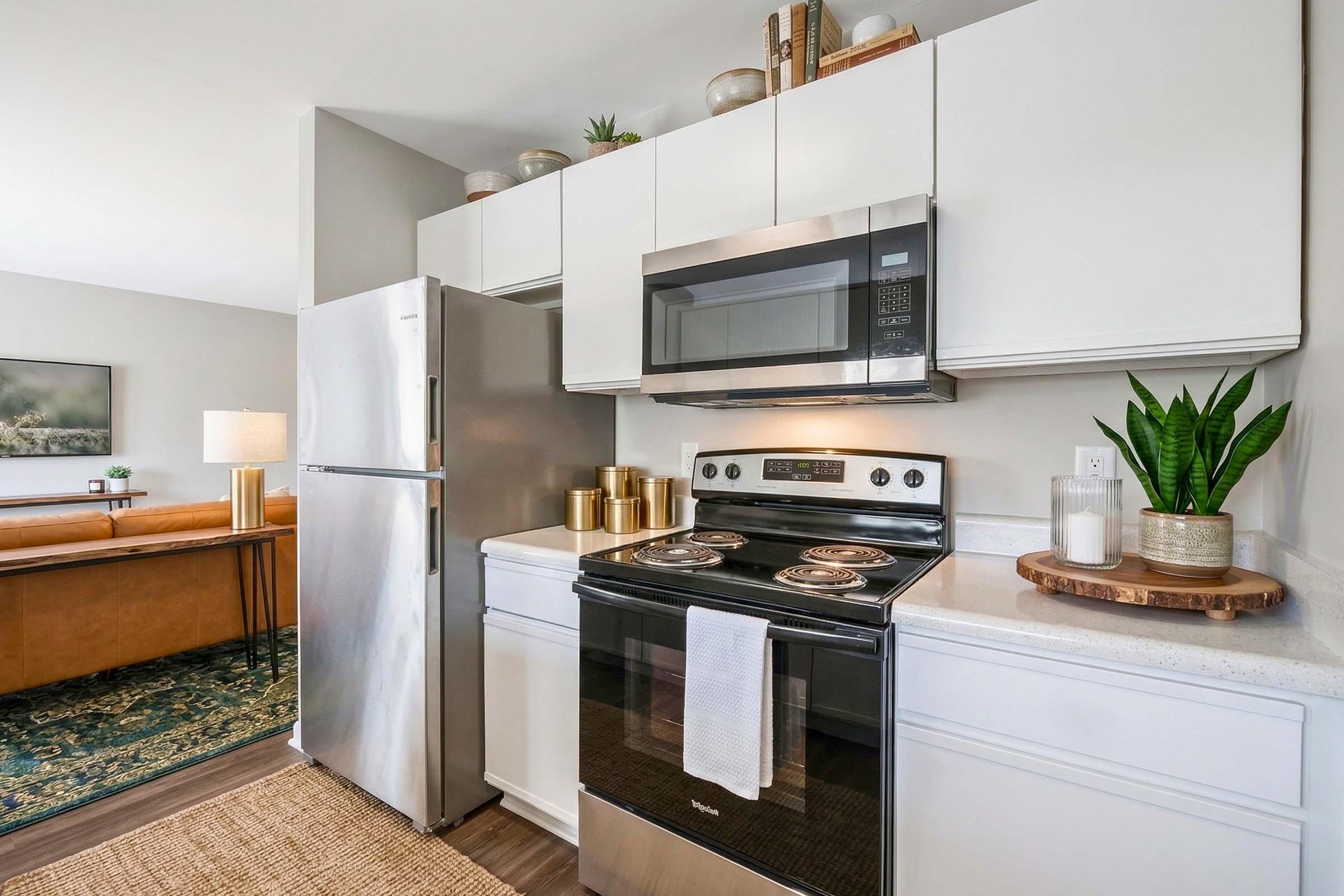 A modern kitchen featuring stainless steel appliances, including a refrigerator and stove. The countertop is clean with a white finish, and there are decorative plants and gold accents. In the background, a cozy living area can be seen with a leather sofa and a lamp, creating a welcoming atmosphere.