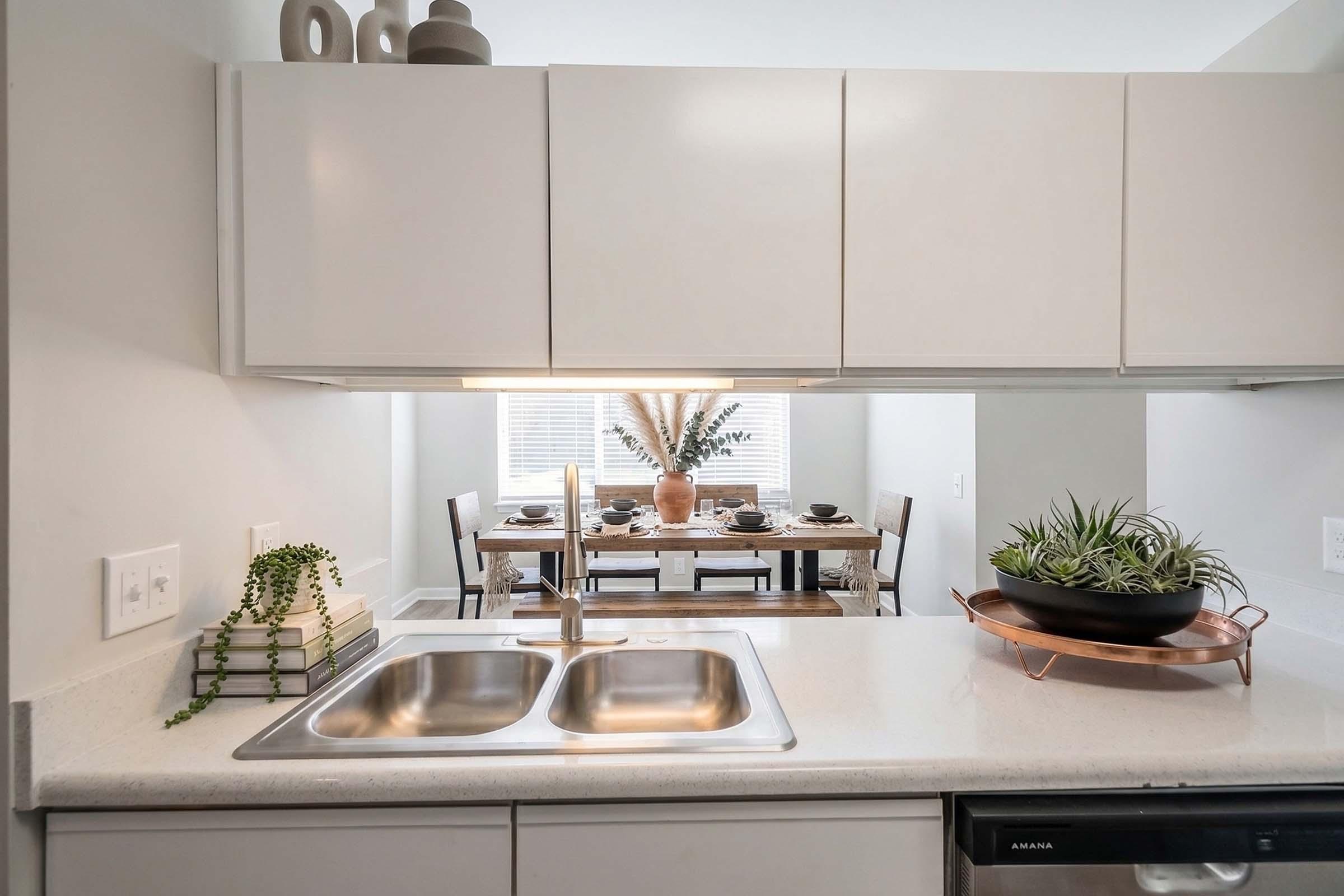 A modern kitchen view with a double sink and minimalist decor. In the background, a dining area features a wooden table set for a meal, adorned with plates and a central vase with greenery. Soft lighting enhances the contemporary aesthetic of the space.