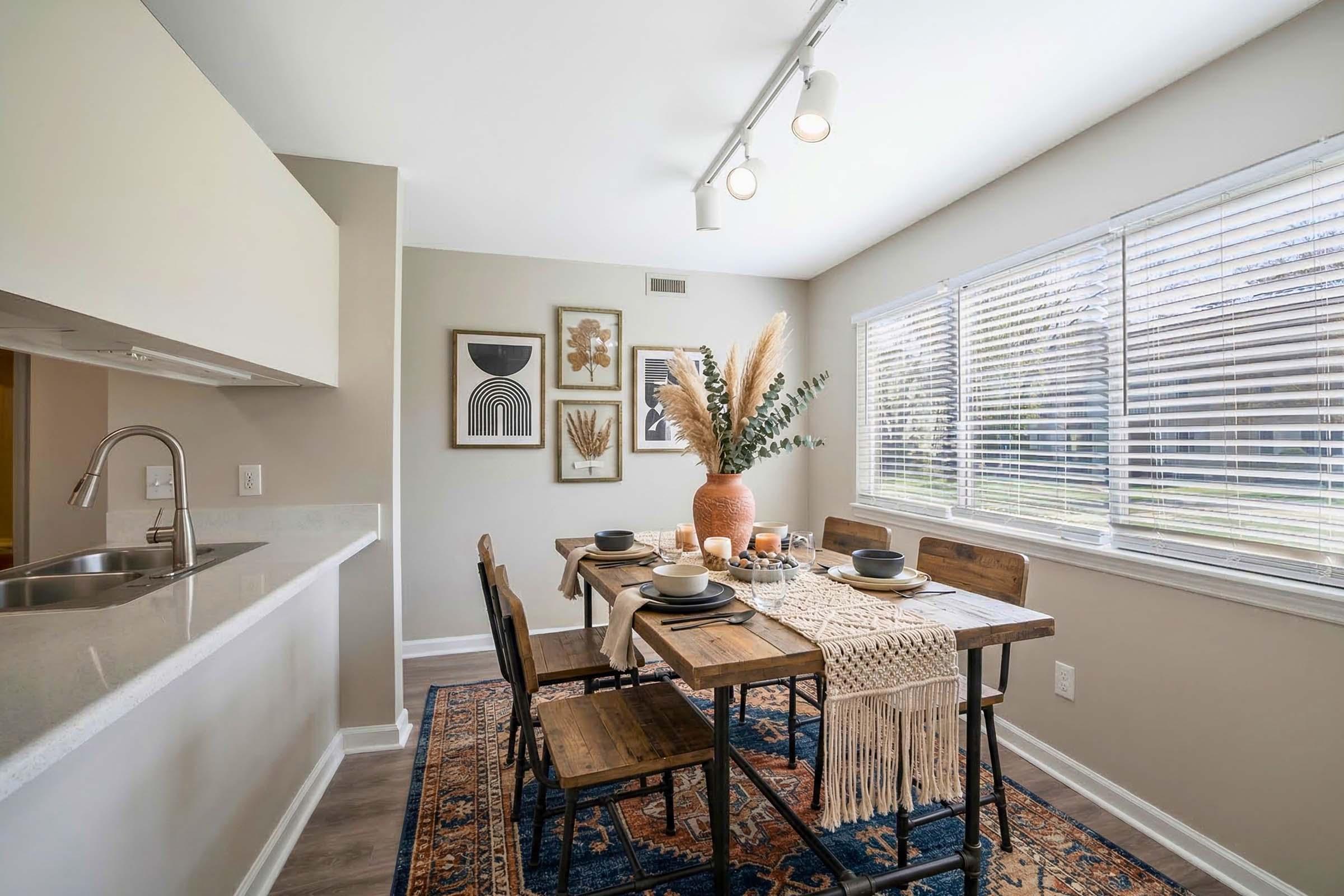 A modern dining area featuring a wooden table set for a meal with dark plates and utensils. The table is adorned with a woven table runner and a decorative vase containing dried plants. Soft light from the large window illuminates the space, and framed artwork hangs on the wall, adding style to the room.