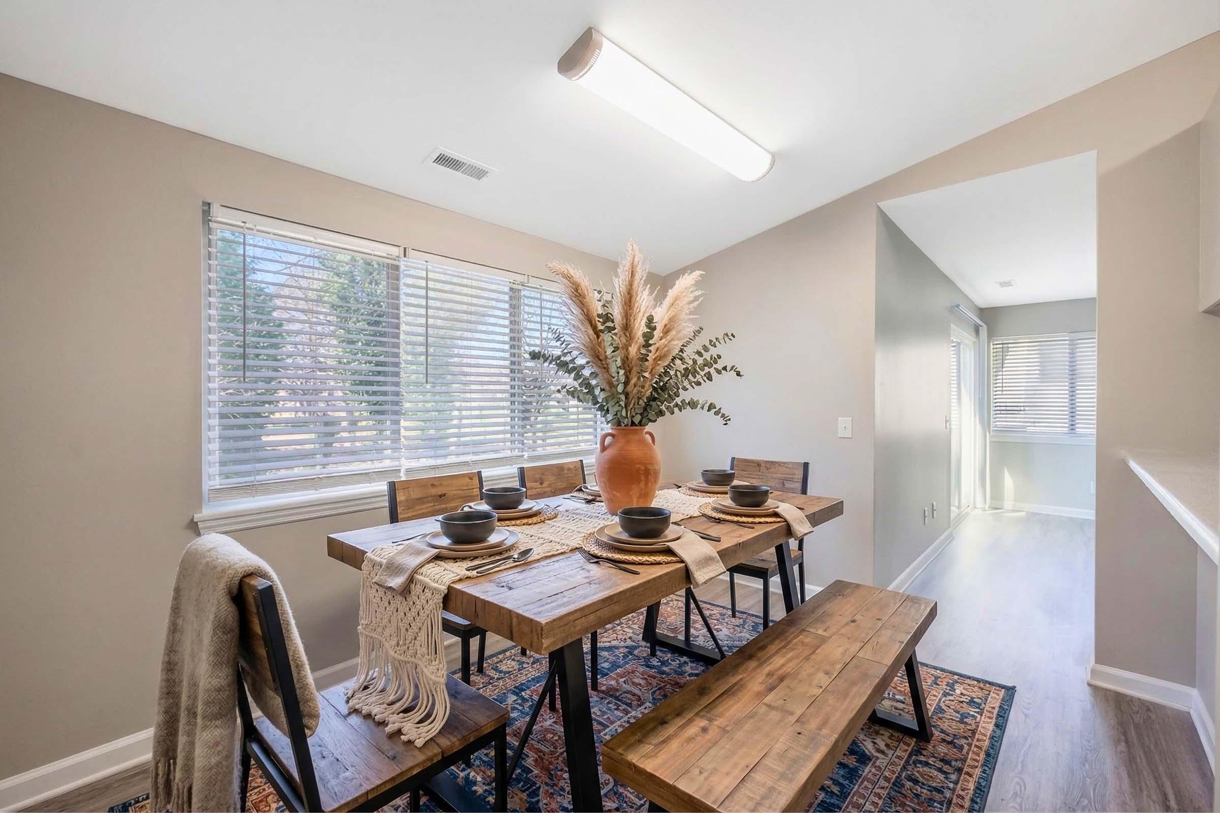 A modern dining area featuring a wooden table with matching benches, set with dark plates and a decorative woven table runner. A large vase filled with dried pampas grass and greenery is the centerpiece. Bright natural light streams in through window blinds, creating a warm, inviting atmosphere.