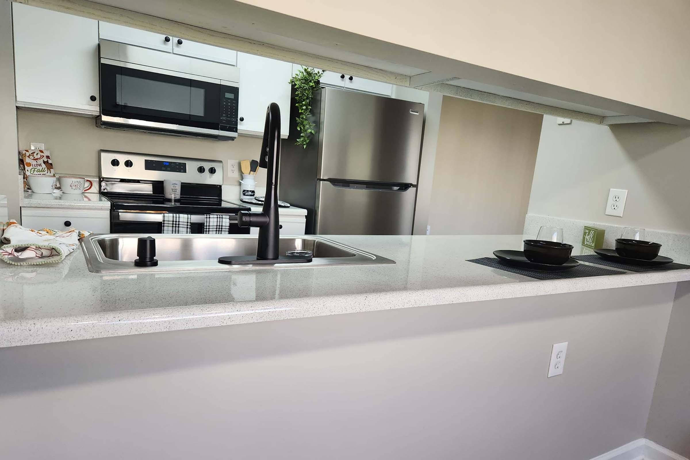A modern kitchen with a view from a countertop. It features a stainless steel sink with a black faucet, two black bowls on a placemat, and a microwave above the stove. The kitchen has white cabinets, a silver refrigerator, and a subtle green plant for decor.