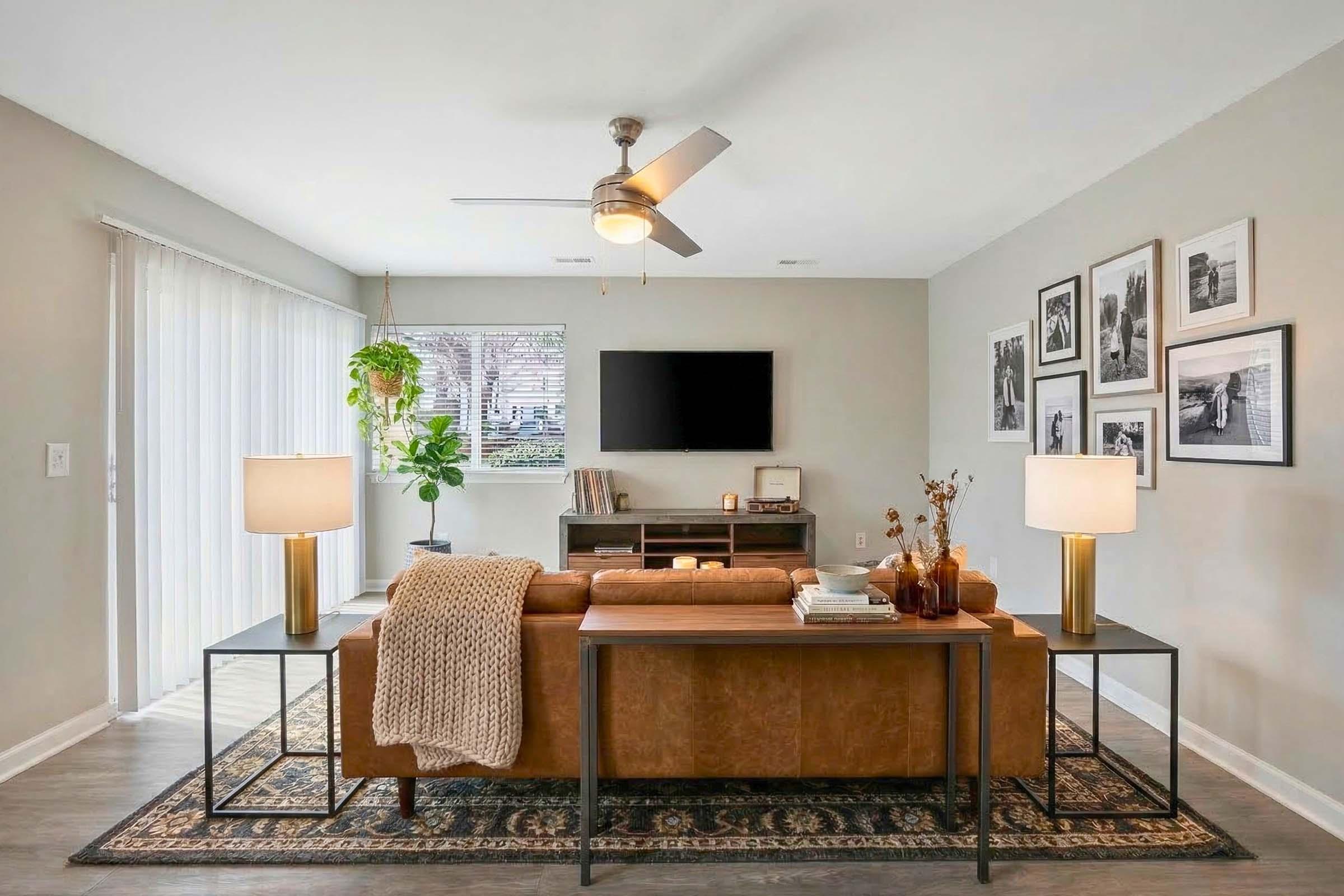 A modern living room featuring a brown leather sofa with a knit throw, two stylish side tables with lamps, and a wooden media console. The walls are adorned with black and white photos, and there's a ceiling fan above. A window allows natural light in, and a plant adds a touch of greenery.