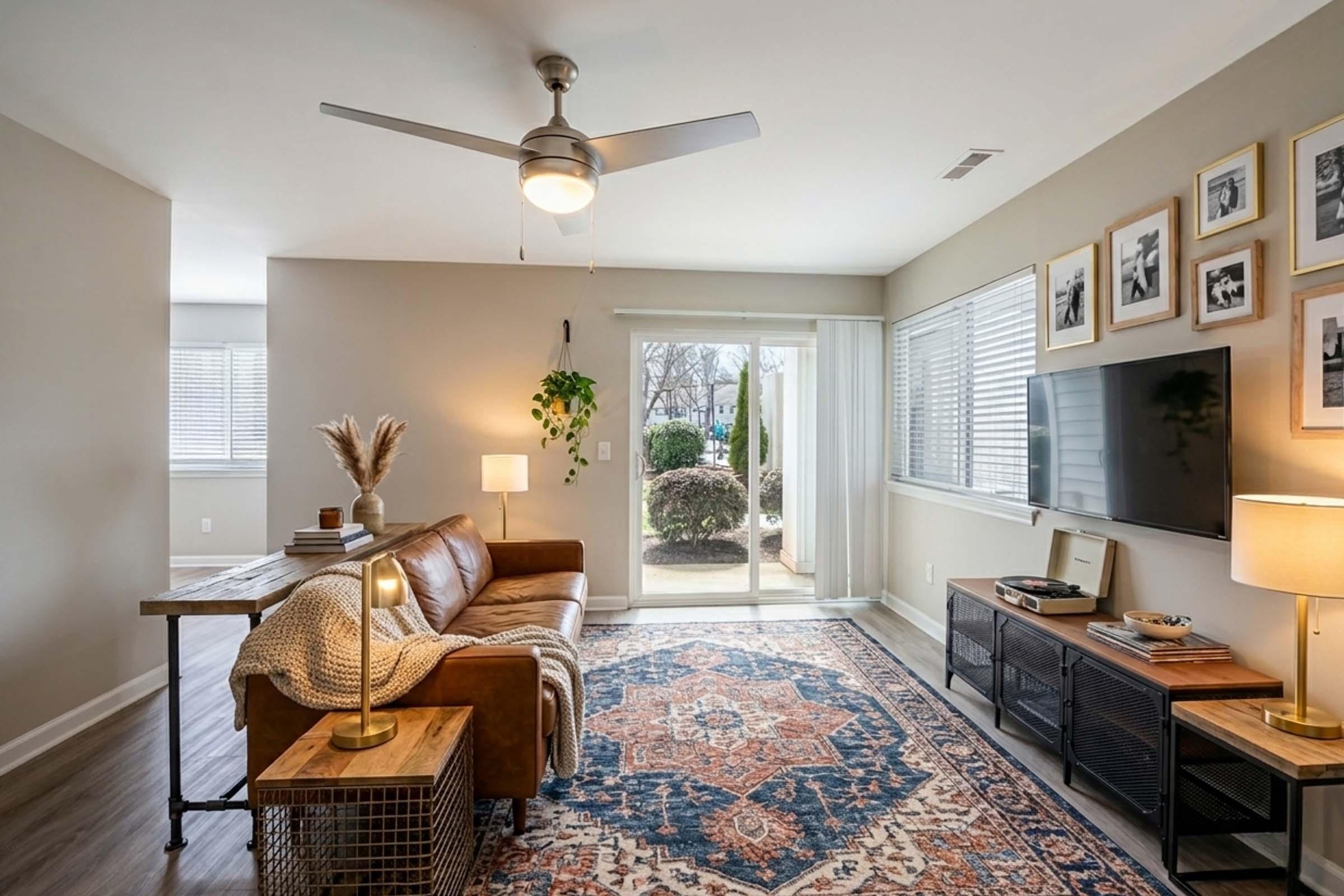 A cozy living room with a brown leather sofa, a wooden table, and a decorative rug. There’s a ceiling fan and a large window with natural light. A flat-screen TV is mounted on the wall, and there are framed pictures. A plant adds greenery, and a stylish lamp stands beside the sofa.