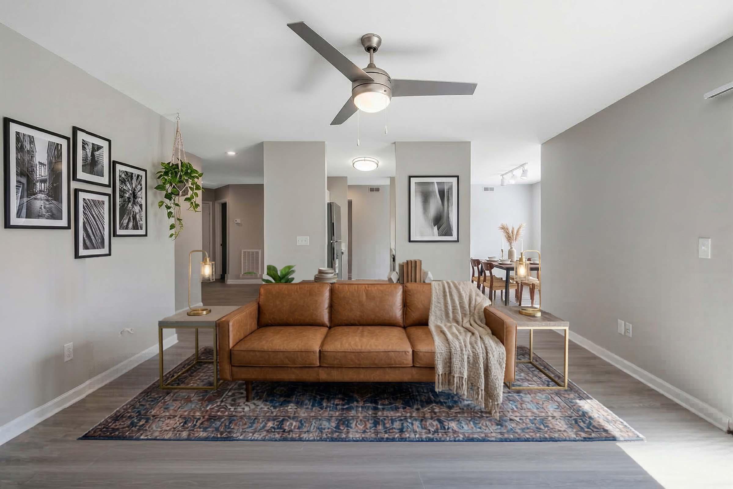 A modern living room featuring a brown leather sofa on a patterned rug, a coffee table with decorative items, and framed artwork on the walls. A ceiling fan is visible above, and the space has a neutral color palette with light wood flooring. Additional furniture and dining area can be seen in the background.
