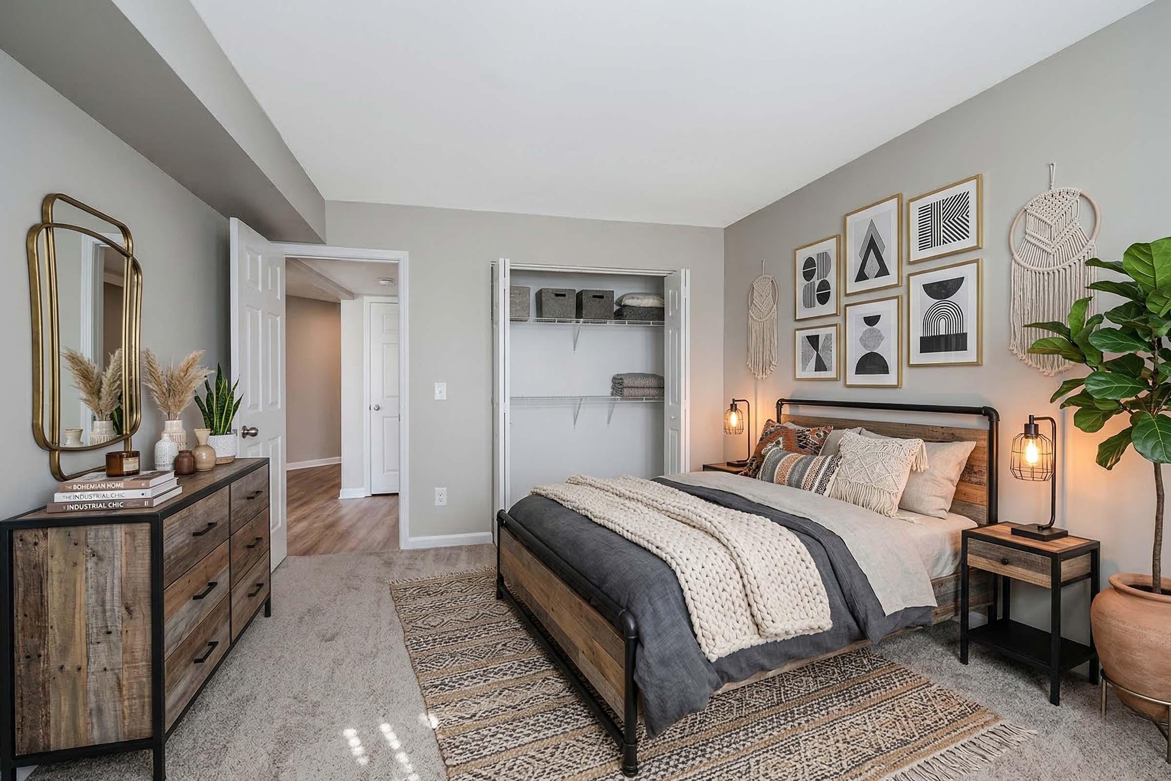 A modern bedroom featuring a bed with neutral bedding, decorative wall art, and a wooden dresser. There are two stylish lamps, a closet without doors, and a large potted plant. The walls are painted light gray, and the room has a cozy, contemporary feel with soft carpeting and natural light.