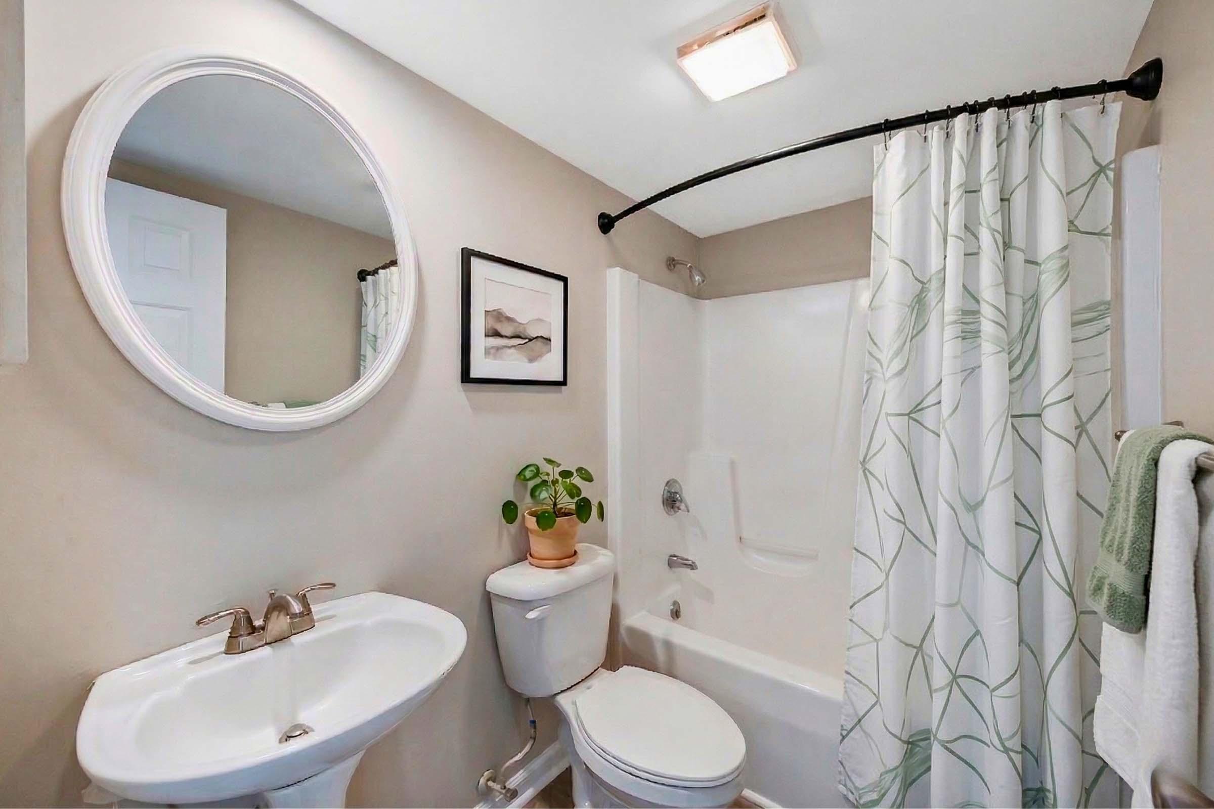 A small, modern bathroom featuring a white bathtub with a shower curtain, a round mirror above a sink, and a potted plant on the floor. The walls are light-colored, and the decor includes a framed picture. A towel hangs on the side of the tub, completing the cozy and clean aesthetic.
