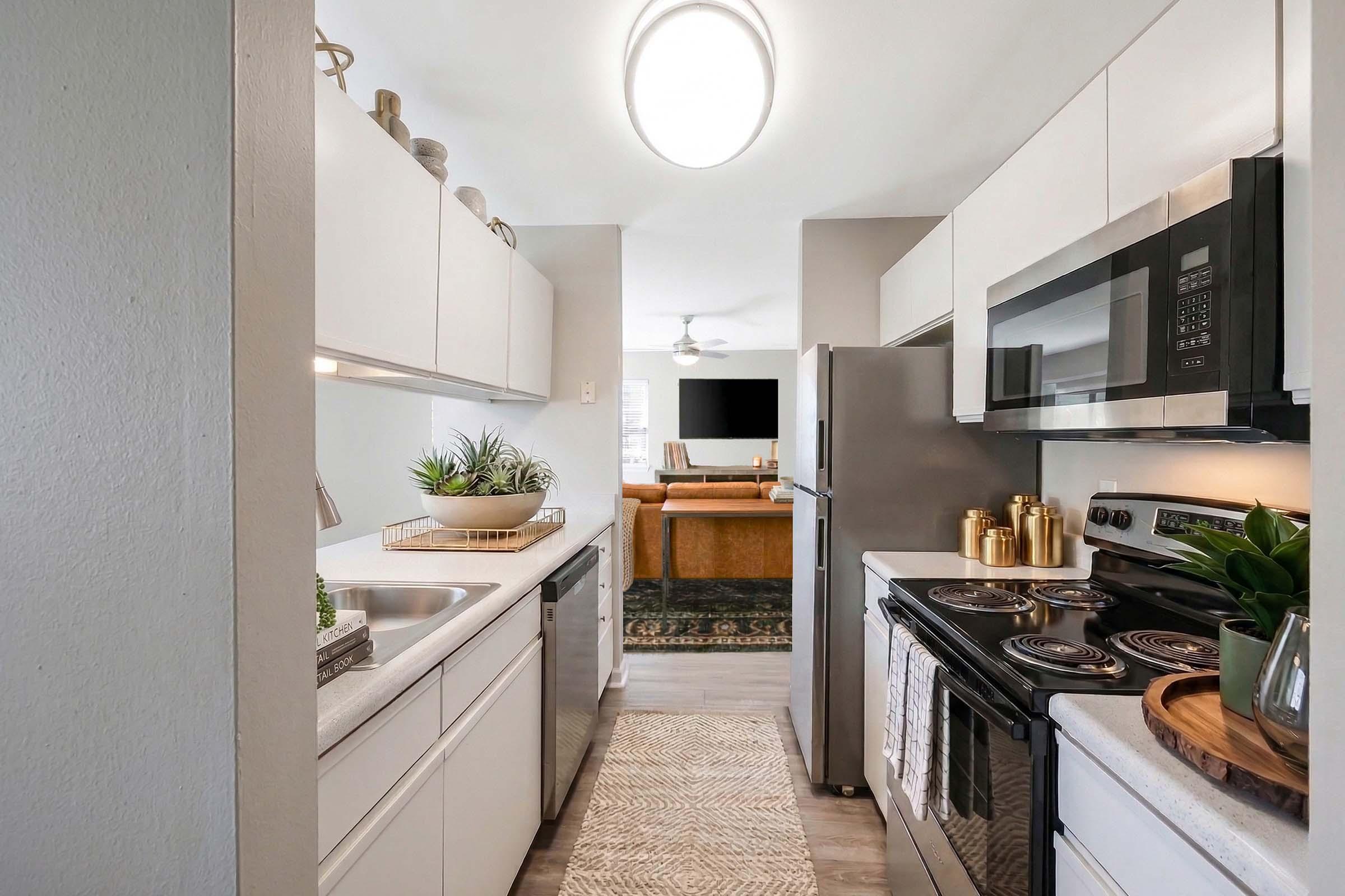 Modern kitchen with white cabinets, stainless steel appliances, and a countertop. Features a double sink with plants, an electric stove, and a microwave. A cozy living room is visible in the background, with a large TV and light-colored furniture. Bright and well-lit space with an inviting atmosphere.