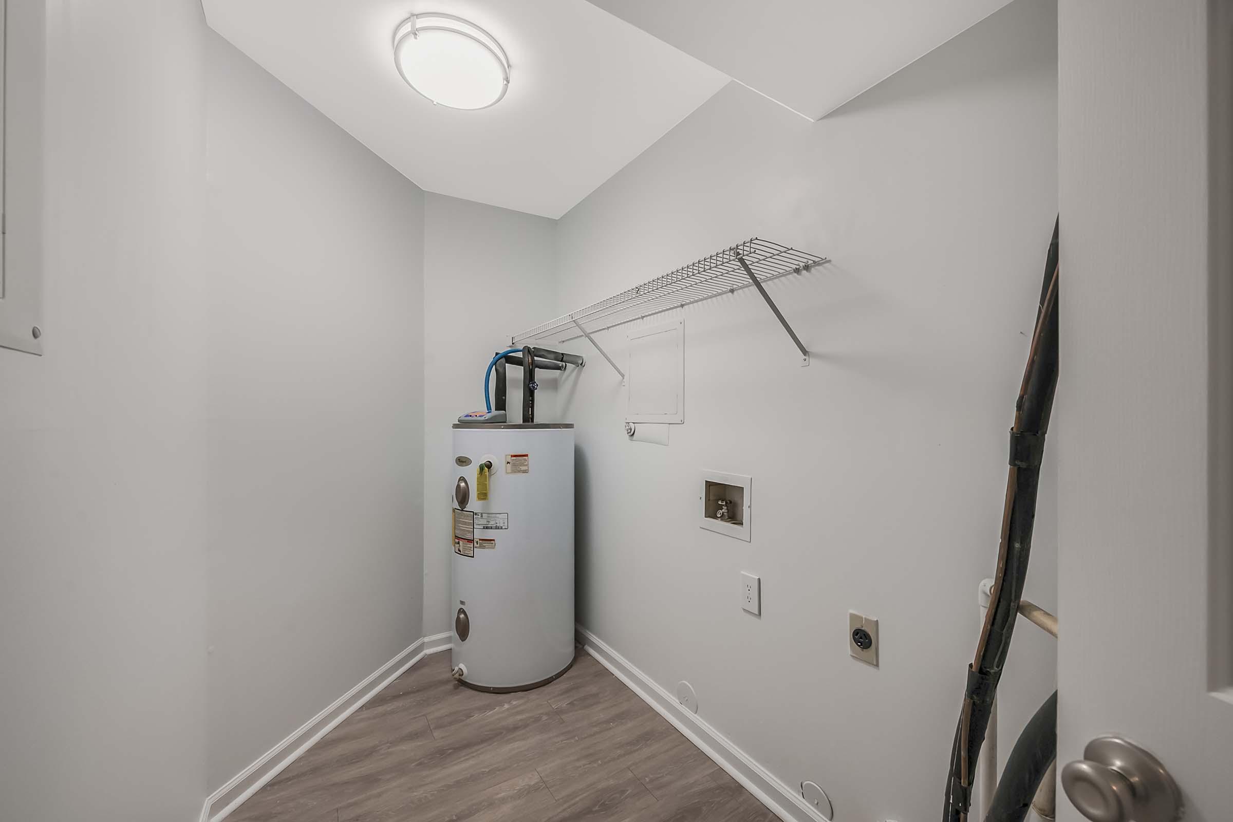 Clean and modern laundry room featuring a white water heater, a wire shelf mounted on the wall, and a light fixture. The walls are painted in a light color and the floor has a wood-like appearance. There are electrical outlets and pipes visible, creating a functional space for laundry activities.