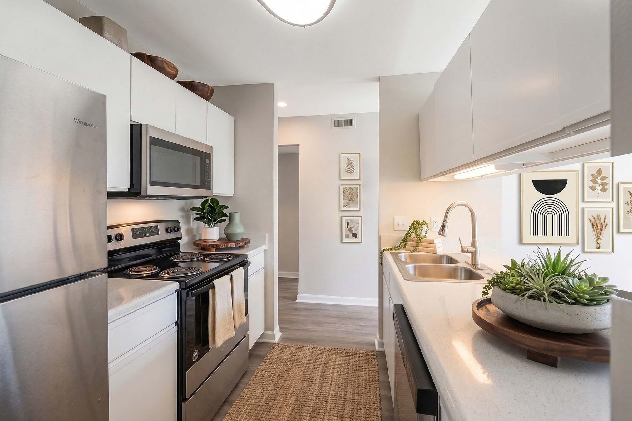 A modern kitchen featuring stainless steel appliances, including a refrigerator, oven, and microwave. The countertops are light-colored with a wooden island table holding a succulent plant. Decorative wall art and house plants add a touch of elegance to the space, which has a bright and airy atmosphere.