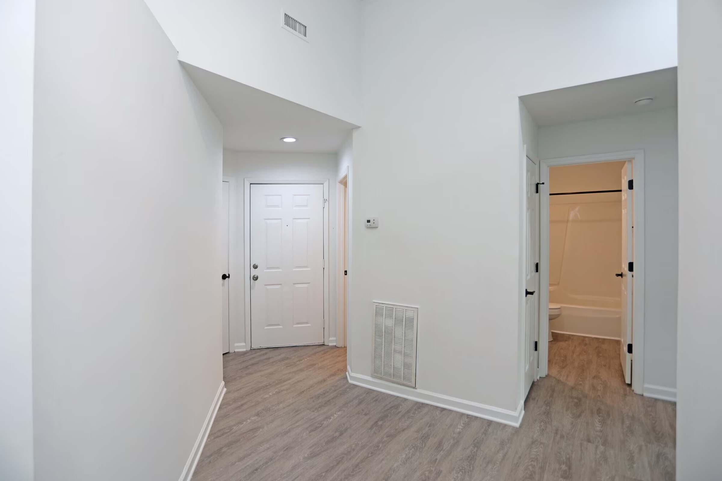 A well-lit hallway with light gray walls and a high ceiling. It features a front door on the left, a bathroom entrance on the right, and a vent on the wall. The floor has a wood-like finish, contributing to a modern and clean aesthetic.