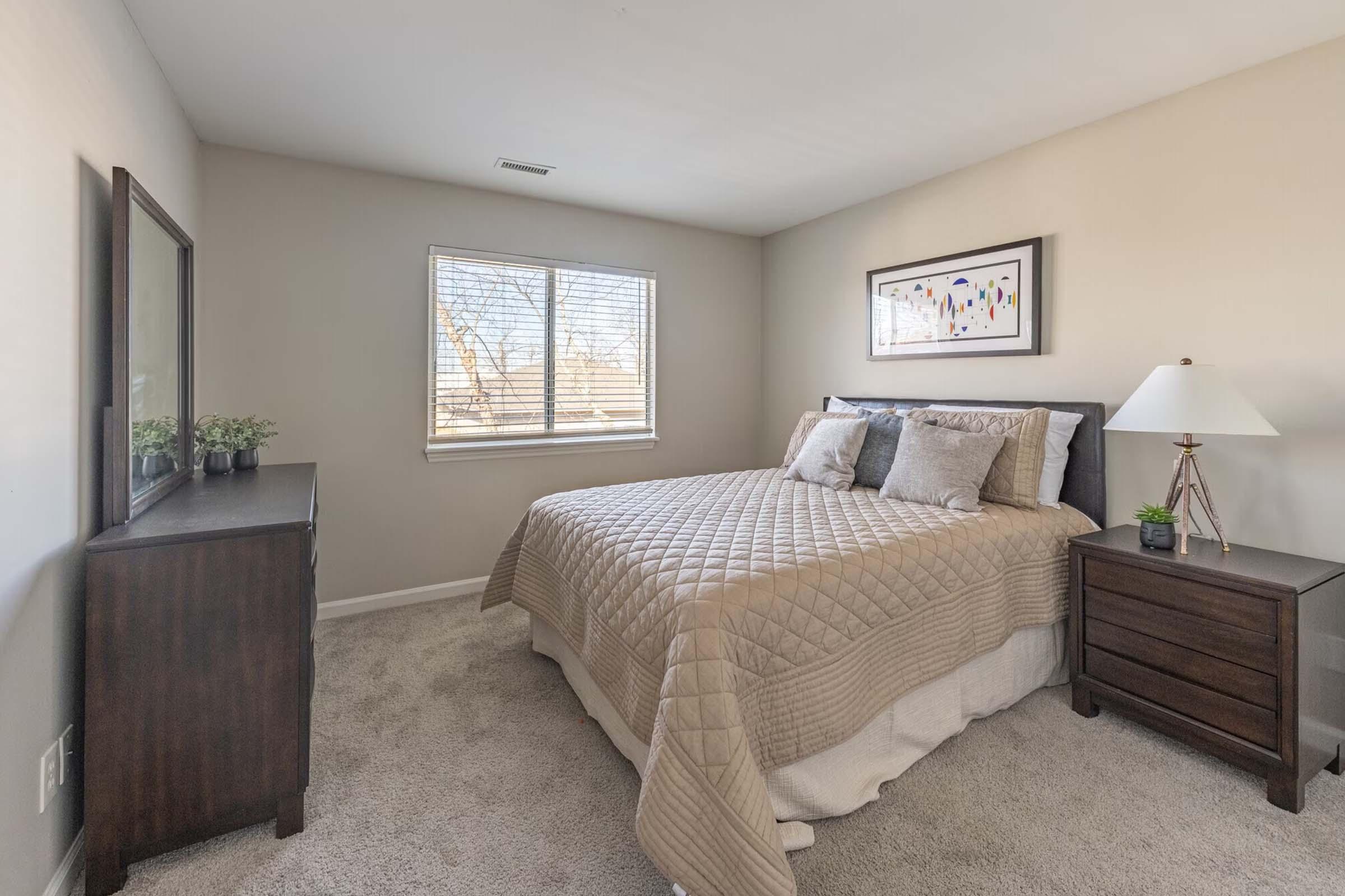 A cozy bedroom featuring a neatly made queen bed with beige bedding, two decorative pillows, and a framed artwork on the wall. A brown dresser and nightstand complete the room, with a lamp on the nightstand and a window allowing natural light to enter, giving a warm and inviting atmosphere.