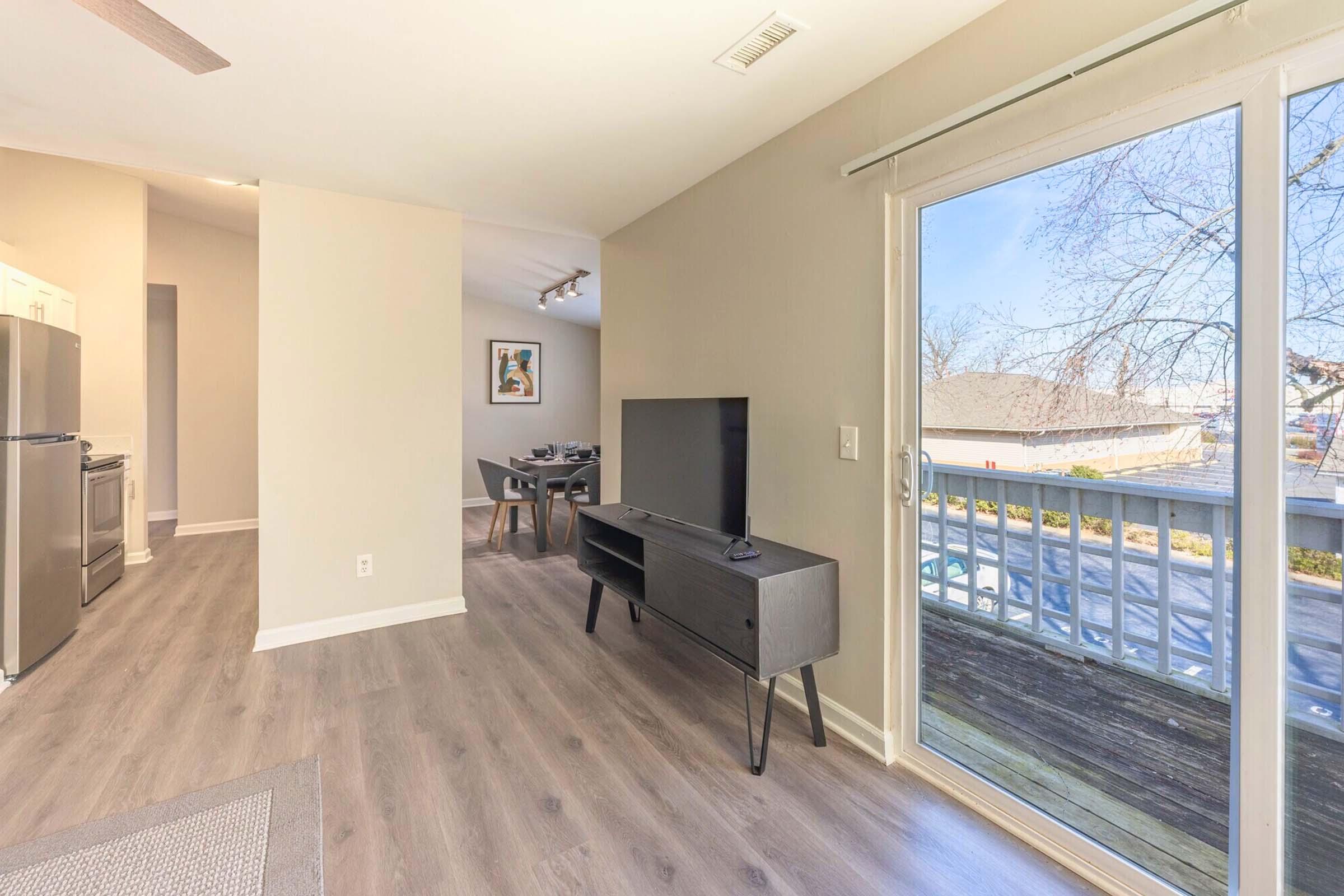 Interior view of a modern living space featuring a dark wooden TV stand with a flat-screen TV, light-colored walls, and large sliding glass doors that open to an outdoor area. The room has contemporary furniture and a dining area visible in the background, with natural light streaming in.