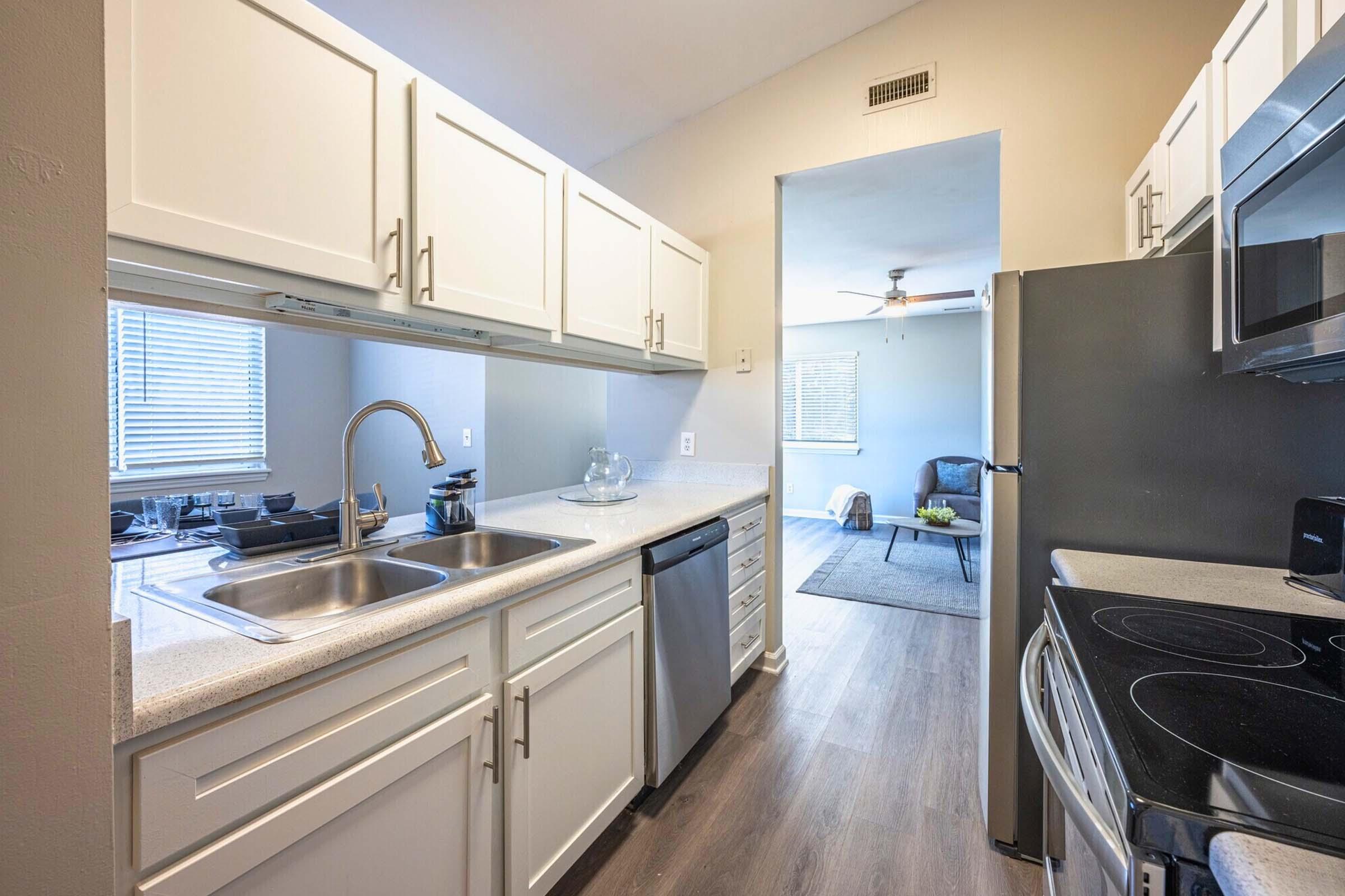 A modern kitchen featuring white cabinets, a double sink, and stainless steel appliances. The view leads to a spacious living area with natural light and a ceiling fan. Wood-style flooring throughout adds a warm touch to the contemporary design.