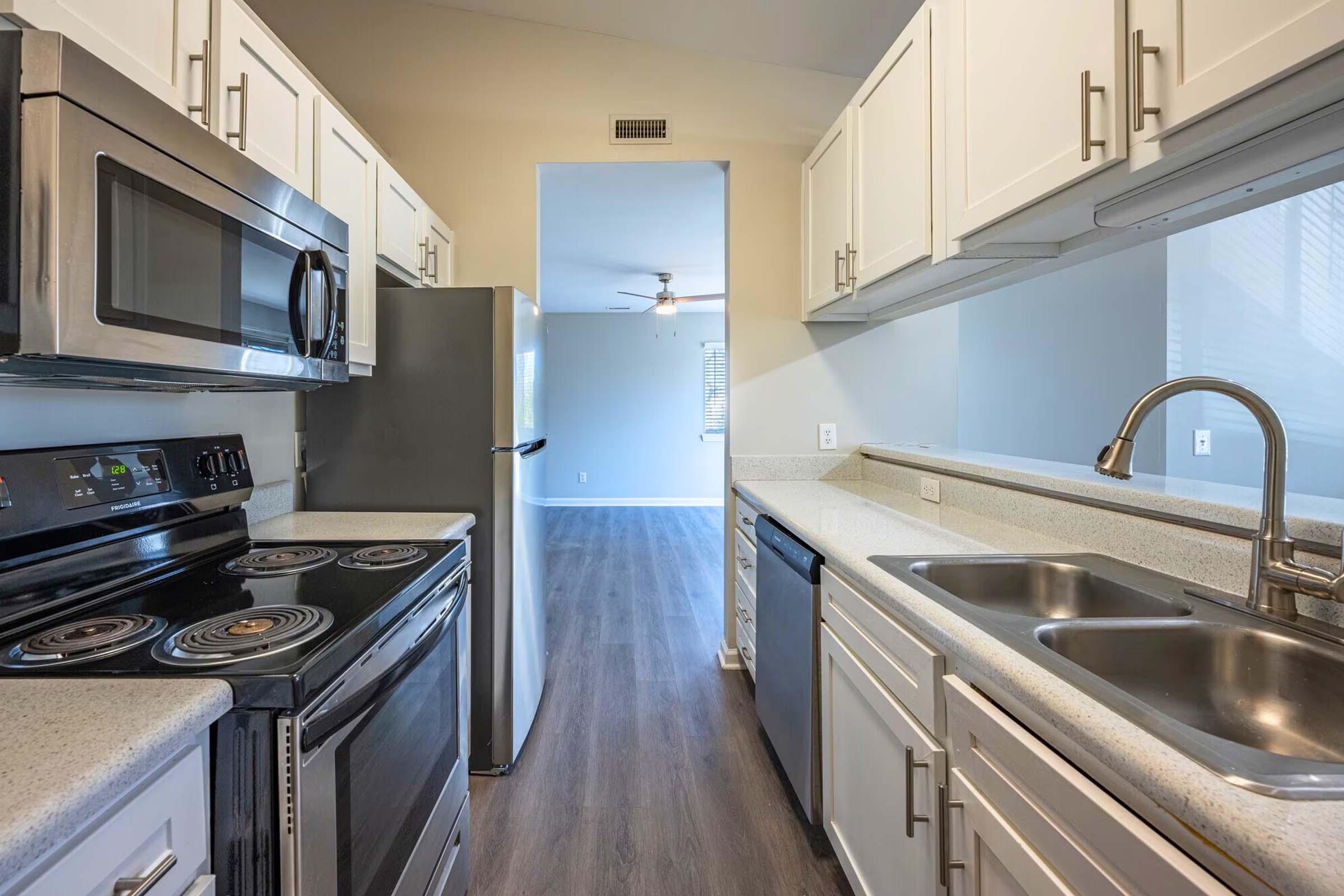 A modern kitchen featuring stainless steel appliances, including a microwave and oven. The countertops are light-colored with a double sink. Kitchen cabinets are white, and there's a view into a spacious living area with wooden flooring and a ceiling fan. Bright, natural light filters in through a window.