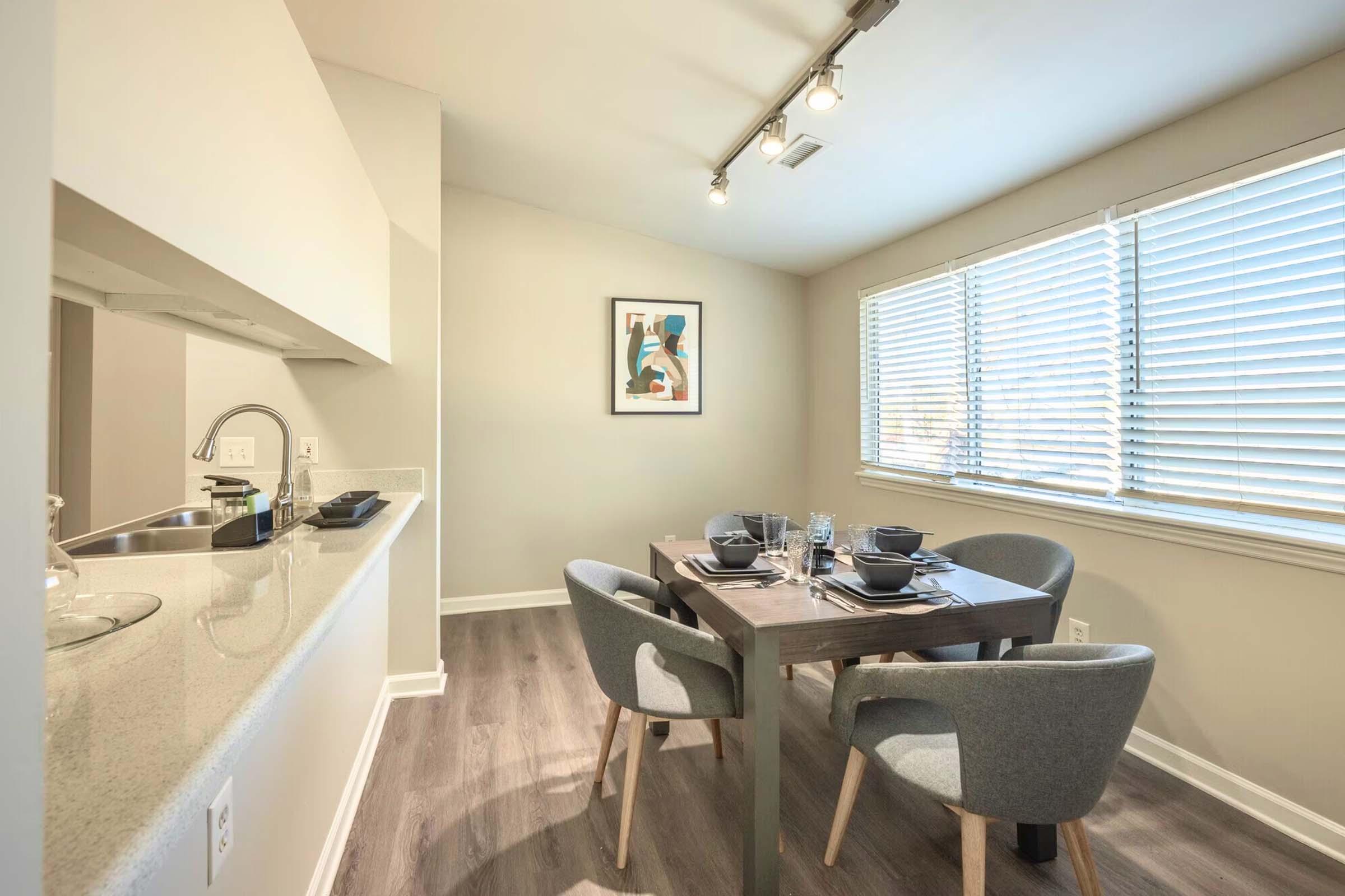 A modern dining area featuring a round table set for four with dark dishes. The room has light wood flooring, gray upholstered chairs, and a decorative wall art piece. Natural light streams in through large windows with blinds. A kitchen area is visible in the background.