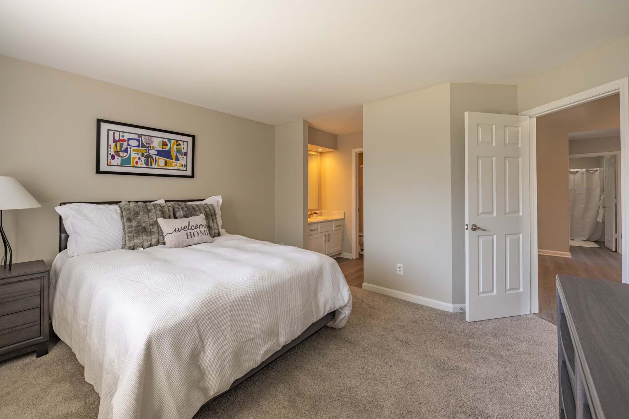A cozy bedroom featuring a queen-sized bed with white linens and decorative pillows, a nightstand with a lamp, and neutral-colored walls. The room has a doorway leading to a bathroom area with soft lighting. There is light brown carpet covering the floor, creating a warm and inviting atmosphere.