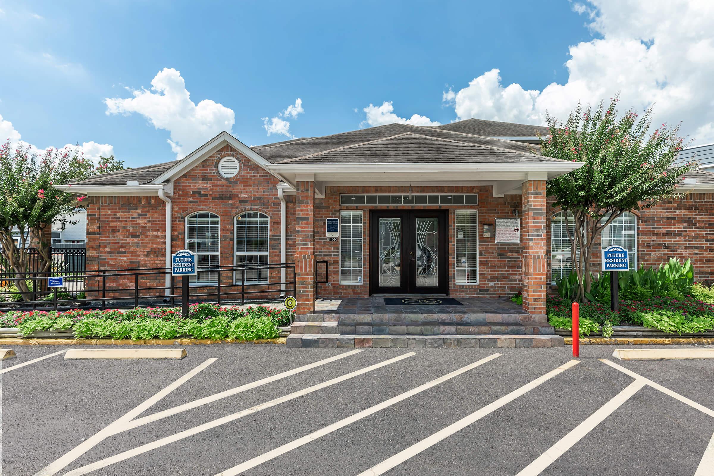 A brick building with large windows and a decorative entrance, surrounded by well-maintained landscaping and flowers. The sky is bright blue with a few clouds. There are clear parking lines and signs indicating parking directions in front of the building.