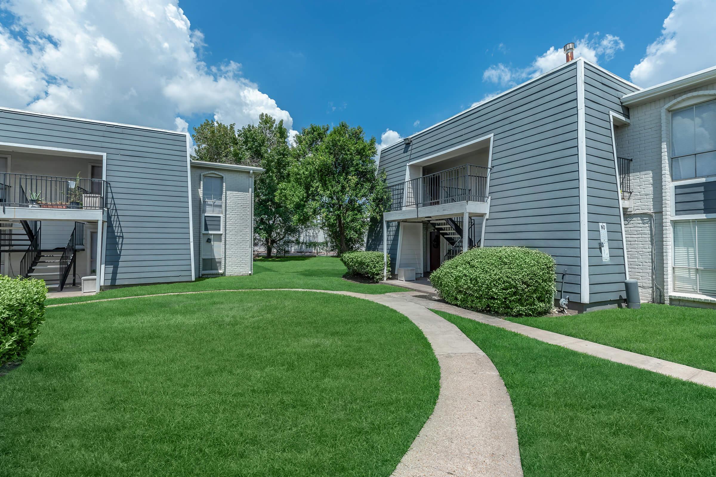 A view of a well-maintained apartment complex featuring two-story gray buildings with balconies, surrounded by lush green lawn and neatly trimmed bushes. A curved pathway leads through the grass, with blue sky and fluffy white clouds visible in the background.