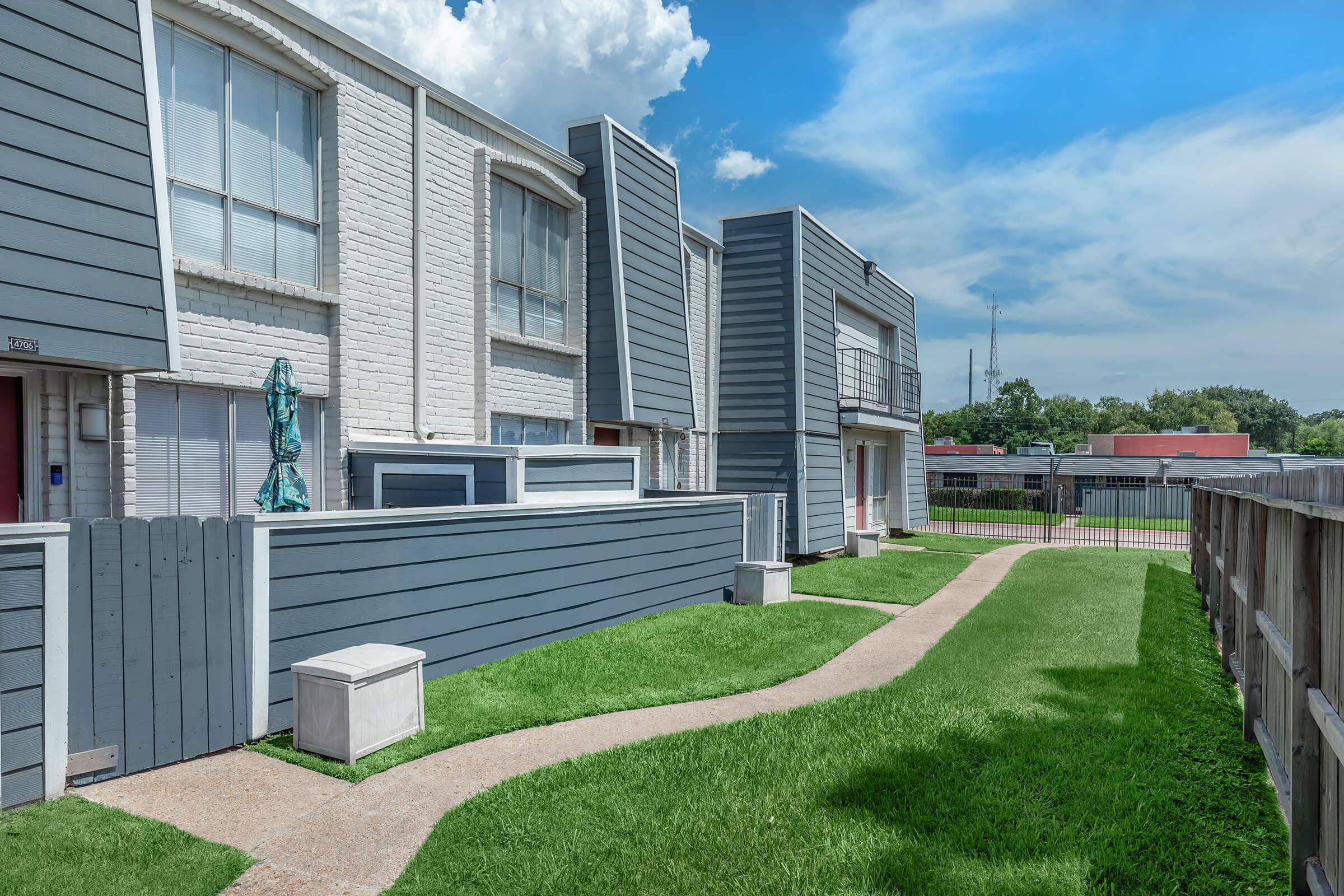 Modern multi-unit buildings with gray siding and white trim, featuring a neatly landscaped lawn and a winding pathway. The sky is partly cloudy, and a fence is visible in the background, separating the area from a nearby parking lot.