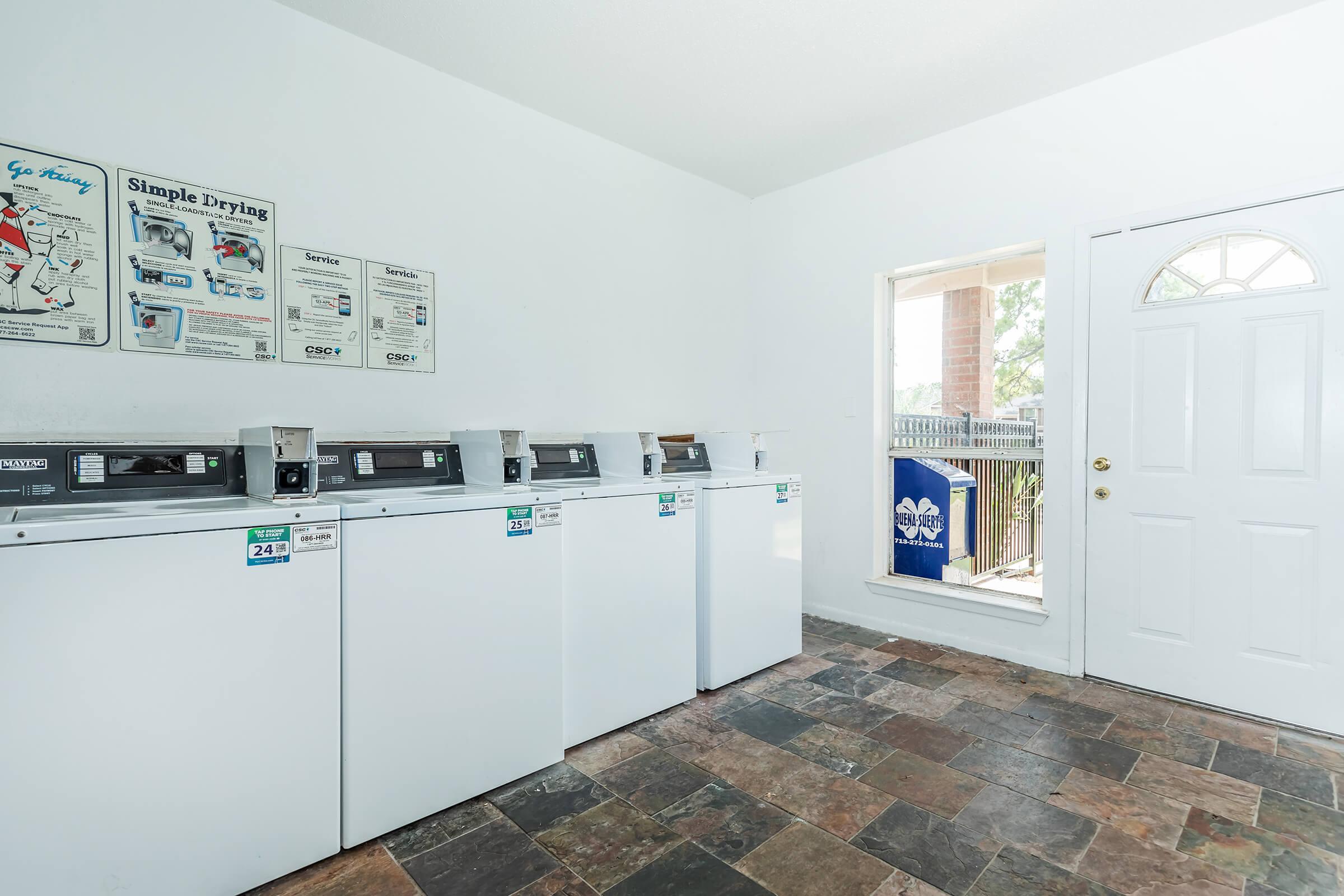 A clean, well-lit laundry room featuring several white washing machines lined up against the wall, with informative posters above. A large window next to a white door lets in natural light, and there's a blue laundry basket outside the window. The flooring is made of dark, slate tiles.