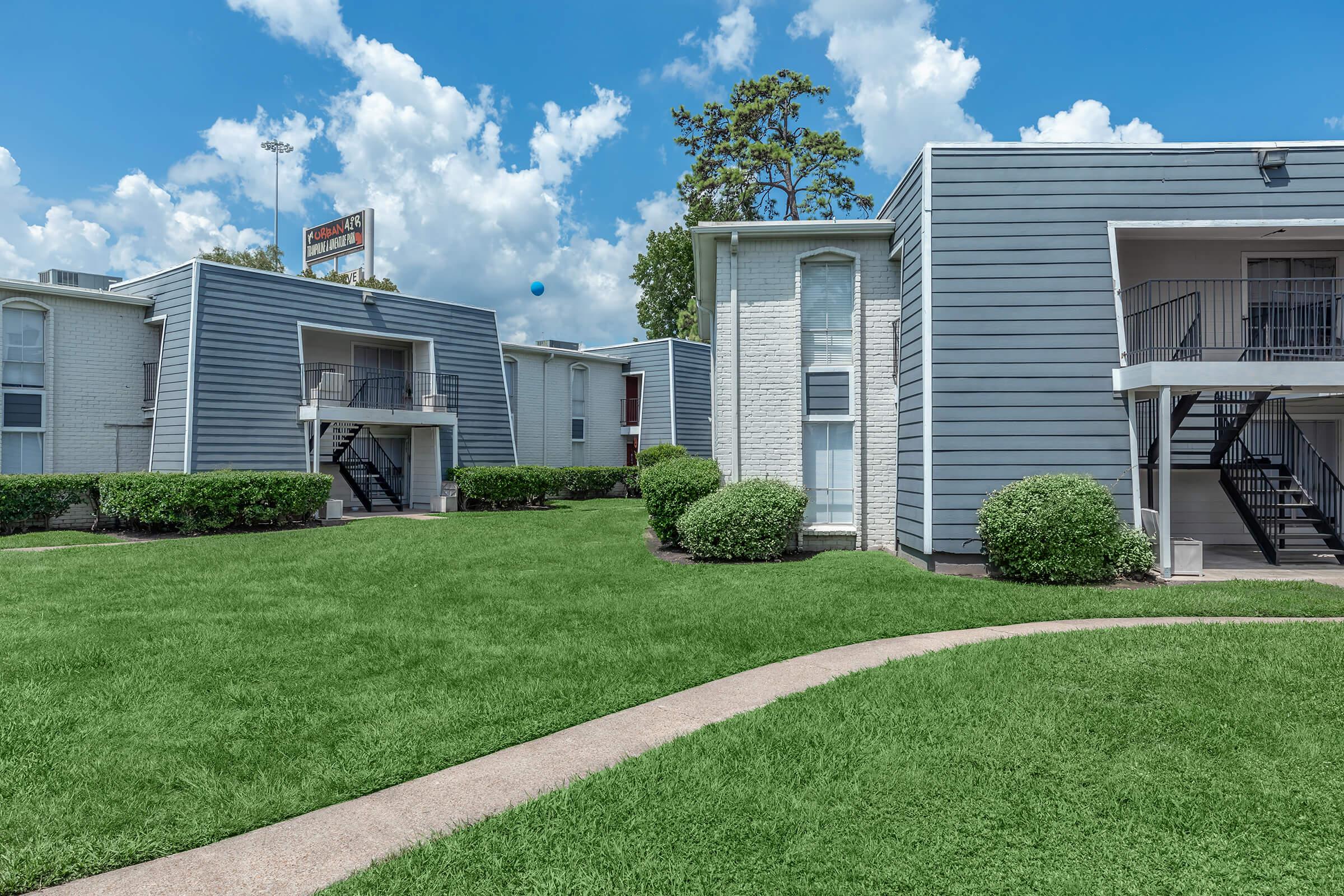 A landscaped area featuring two gray apartment buildings with staircases, surrounded by neatly trimmed shrubs and a grassy lawn under a bright blue sky with fluffy clouds. A sign is visible in the background, indicating the presence of the apartment complex.