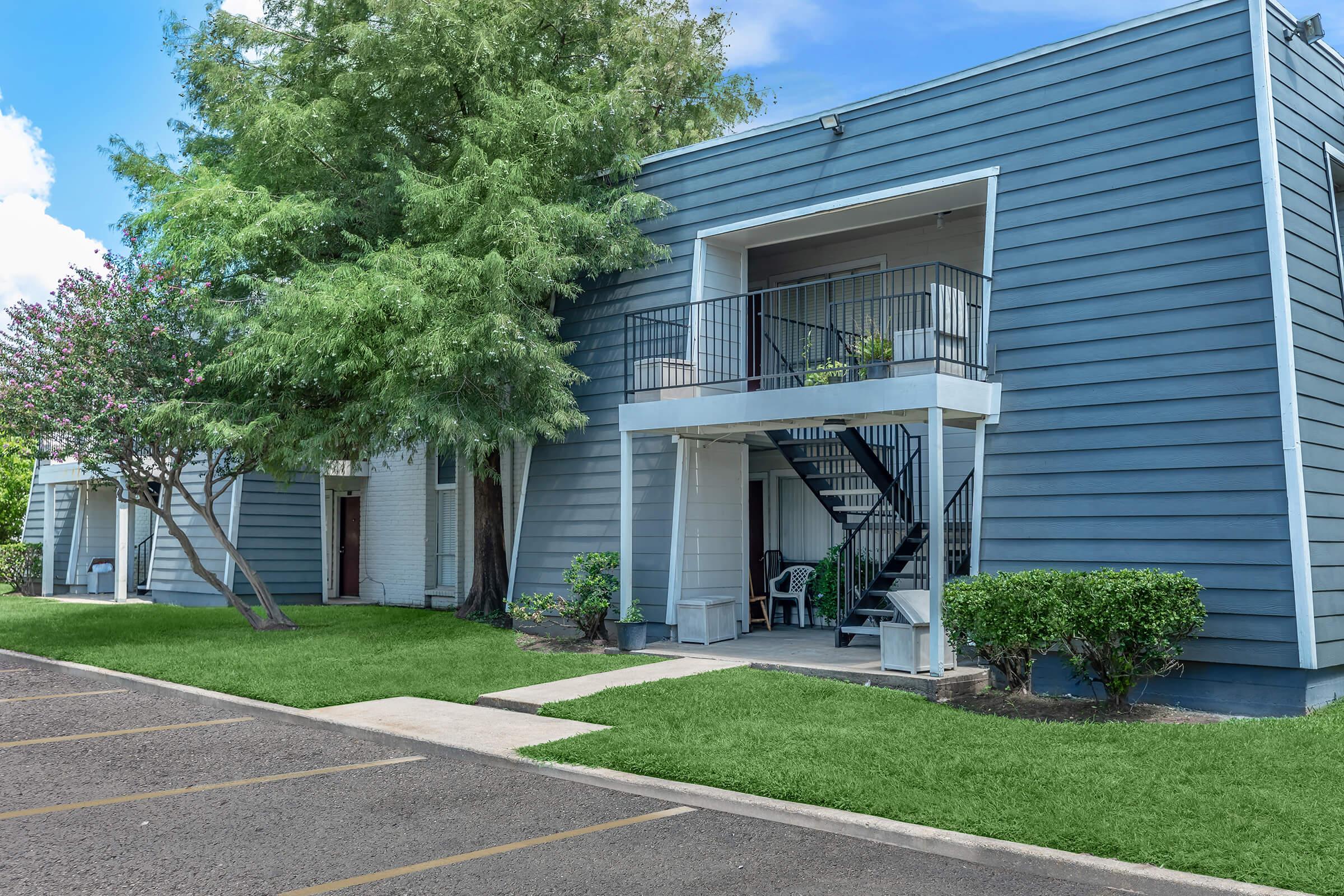 Two-story apartment building with blue siding, featuring a staircase leading to a balcony. Lush green grass surrounds the entrance, and a few small trees and shrubs are present. Clear blue sky in the background, with some fluffy clouds. A parking lot is visible in the foreground.