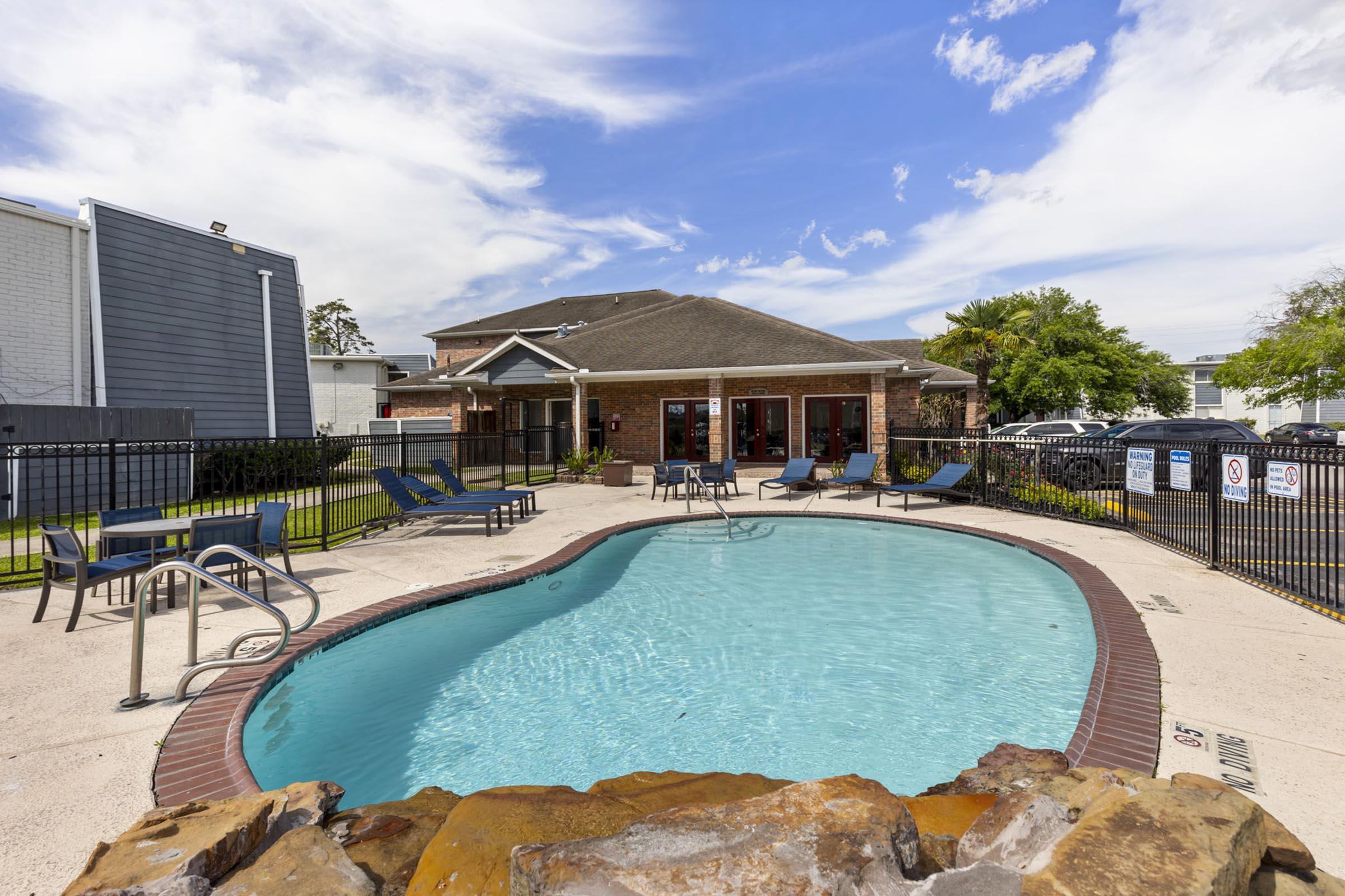 A sunny pool area featuring a clear blue swimming pool with surrounding lounge chairs. In the background, a brick building with large windows and a canopy can be seen, with trees and a blue sky providing a pleasant atmosphere. Fences and parking spots are visible nearby.