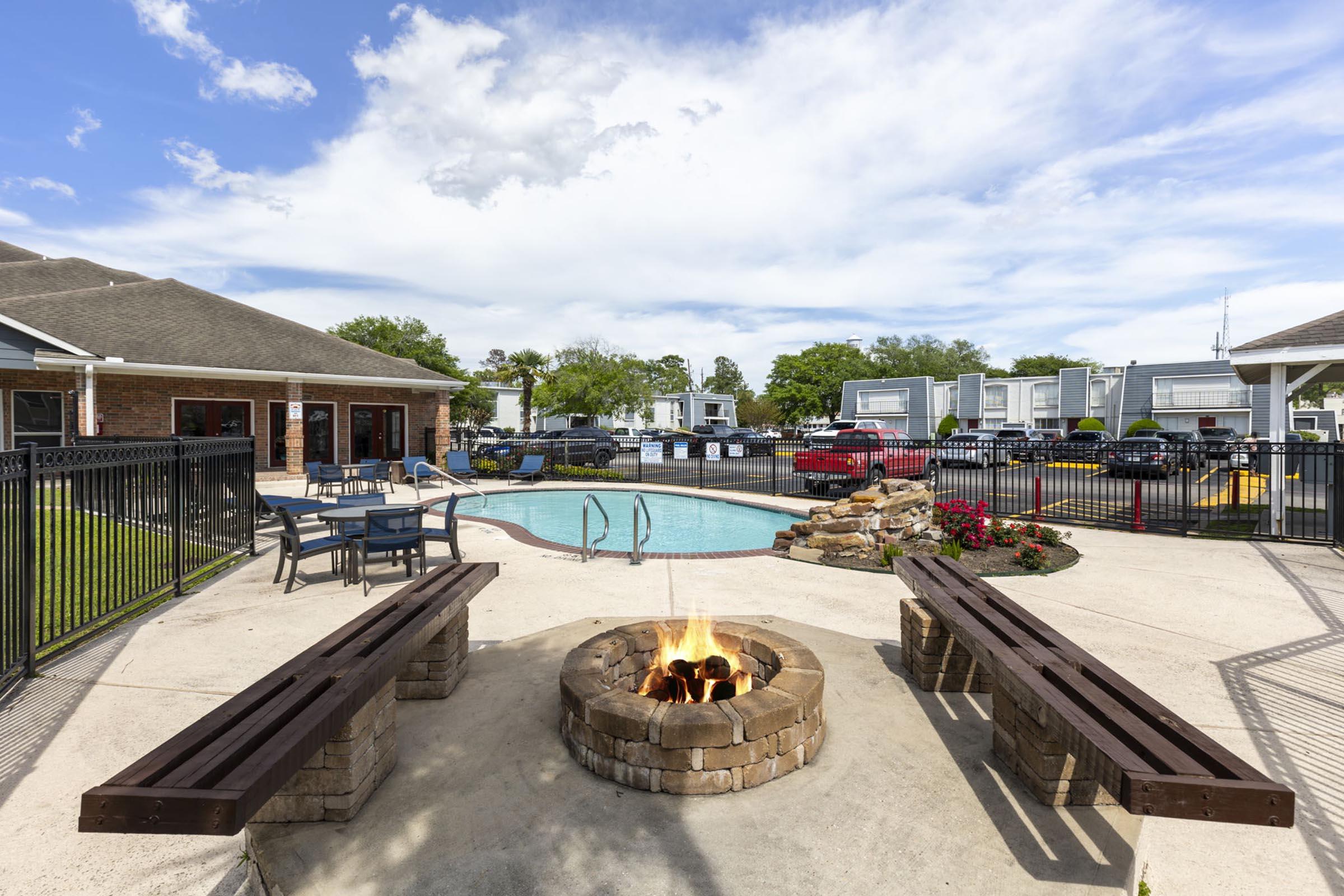 A cozy outdoor area featuring a stone fire pit surrounded by wooden benches, with a swimming pool and lounge chairs nearby. Lush green grass and colorful flowers enhance the atmosphere, while a clubhouse is visible in the background under a partly cloudy sky.