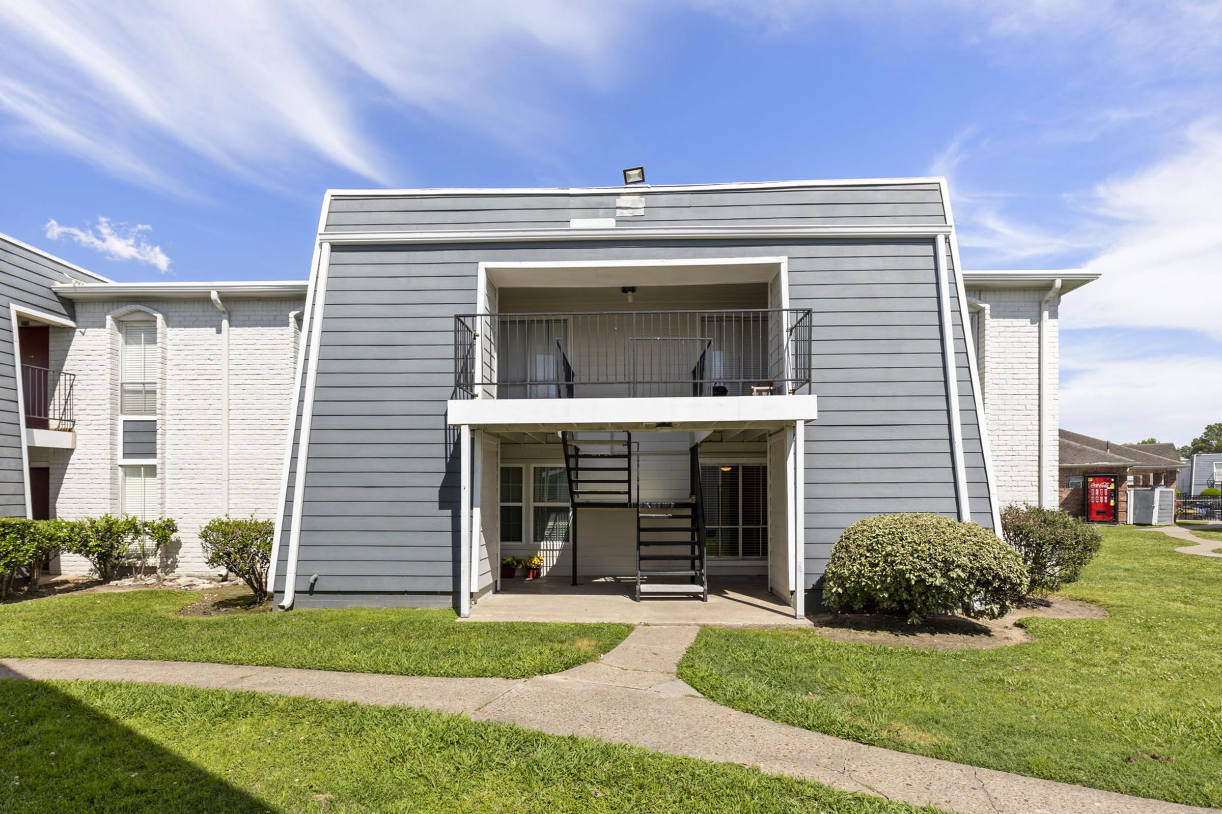 Two-story apartment building with a gray exterior featuring a staircase leading to a balcony. The ground floor has a small patio area, and the surrounding landscape includes neatly manicured grass and shrubs. The sky is blue with a few clouds.