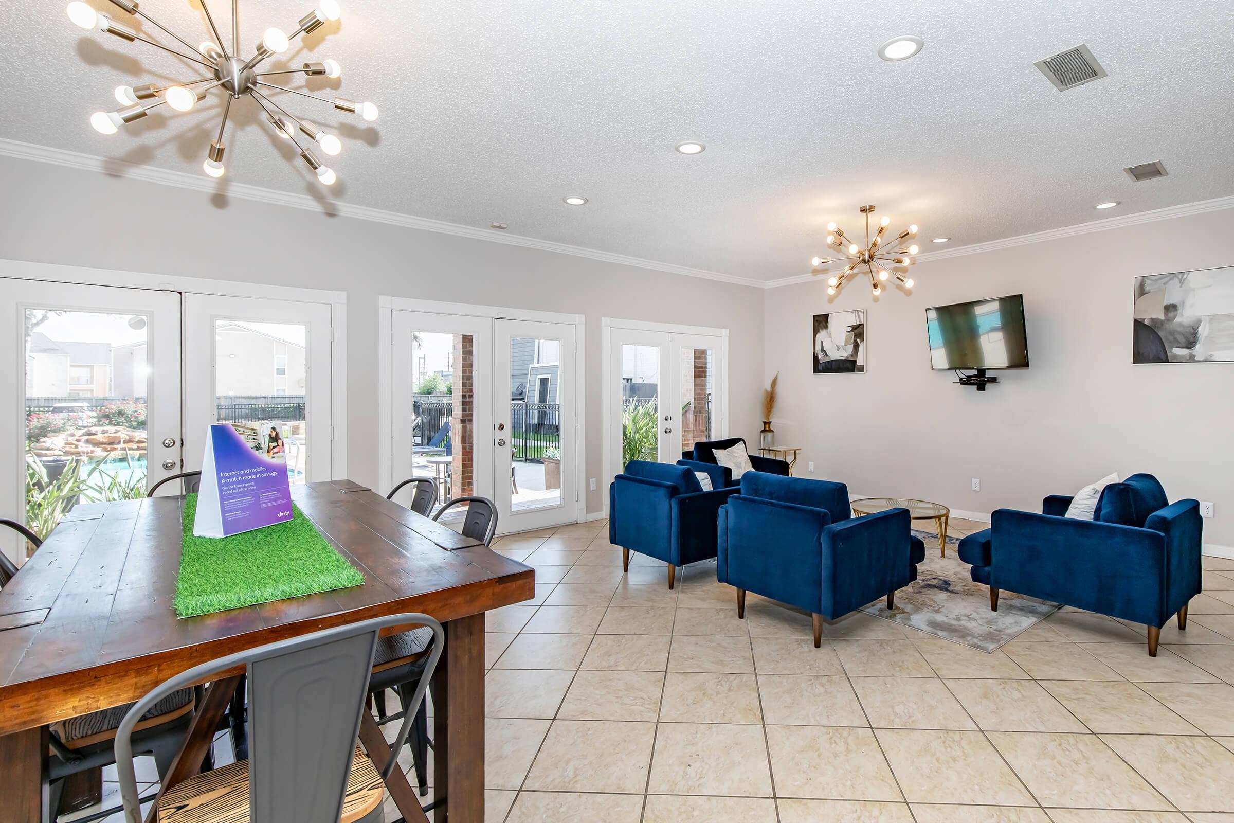 A modern living space featuring a wooden dining table with metal chairs, blue velvet armchairs, and a TV mounted on the wall. Brightly lit with stylish light fixtures, the room has large windows that open to a patio area, creating an inviting atmosphere. Neutral-colored walls and tiled flooring complete the decor.