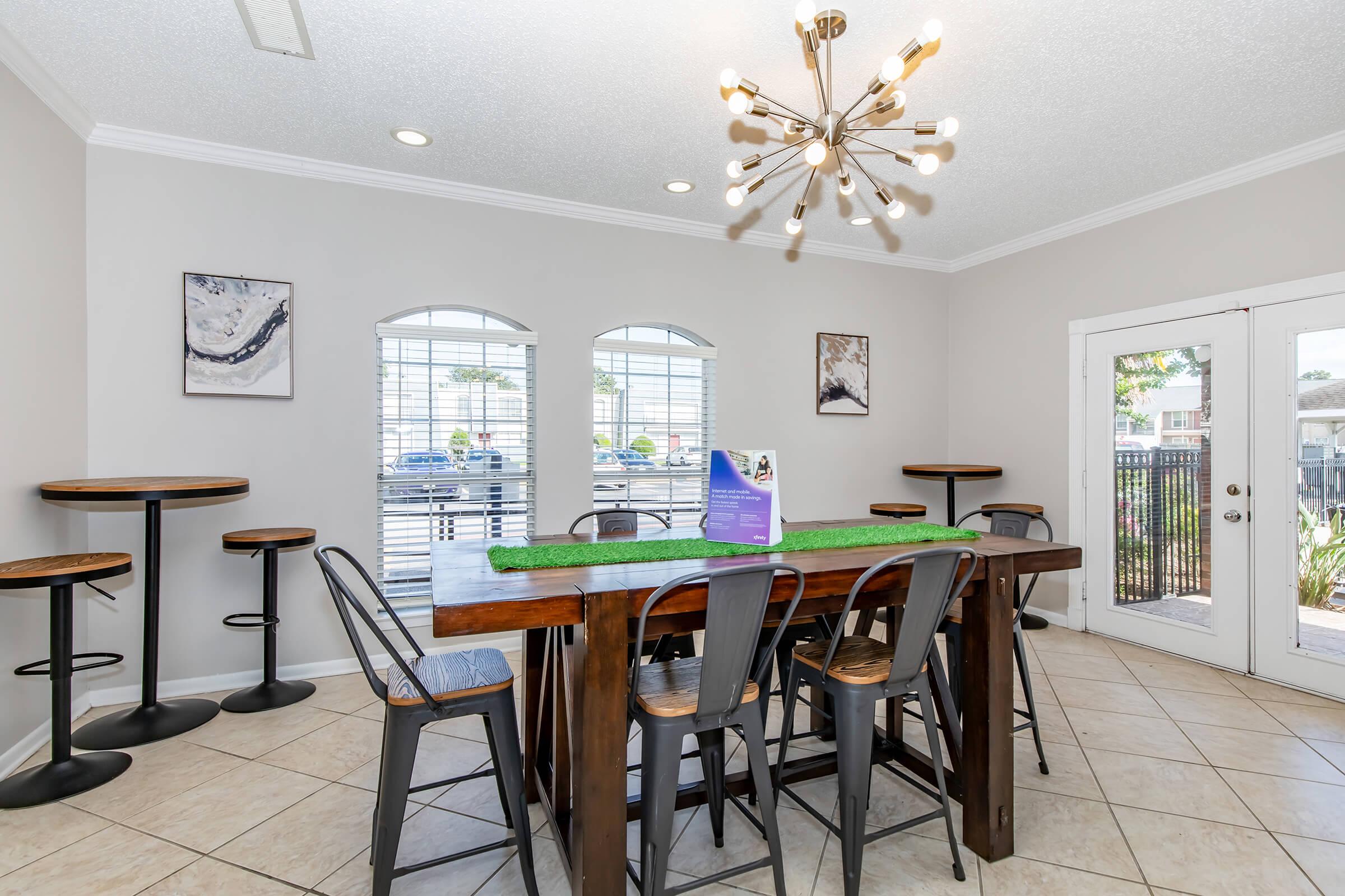 A modern dining area featuring a large wooden table with high stools, surrounded by large windows with blinds. There is a decorative light fixture hanging from the ceiling and a small green tabletop centerpiece. The floor is tiled, and sliding glass doors lead to an outdoor view.