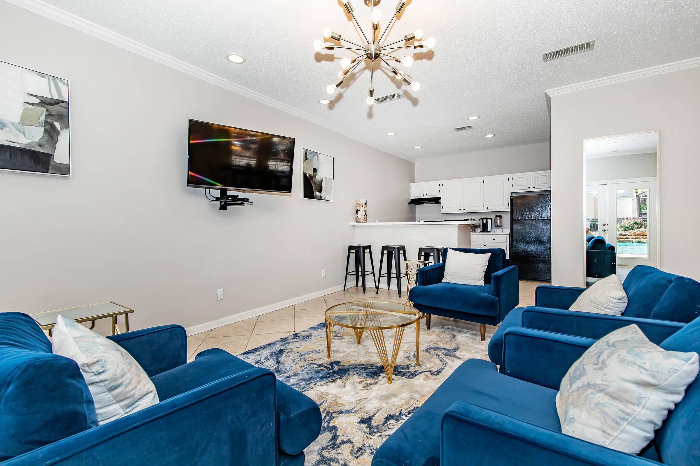 A modern living room featuring blue upholstered chairs arranged around a circular glass coffee table on a patterned rug. A flat-screen TV is mounted on the wall, and a kitchen area with white cabinets and bar stools is visible in the background. The ceiling is adorned with a unique chandelier.