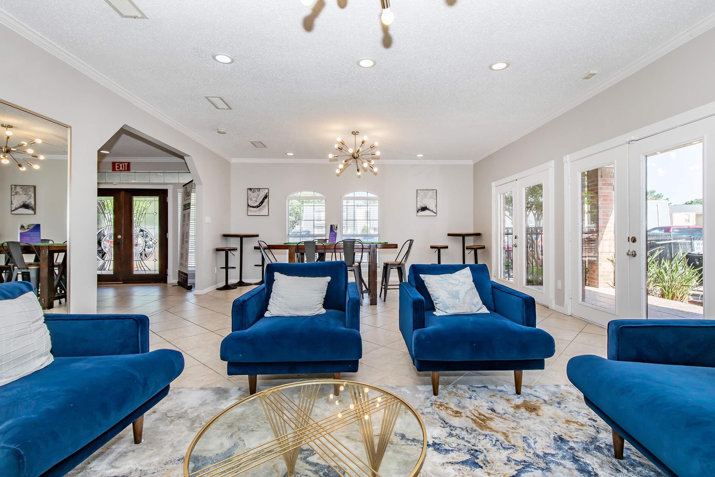 A modern living area featuring blue armchairs with white pillows, a round glass coffee table, and light-colored tiled flooring. The space has a bright atmosphere with large windows, a dining area visible in the background, and artistic wall decor. Stylish lighting fixtures add to the contemporary design.