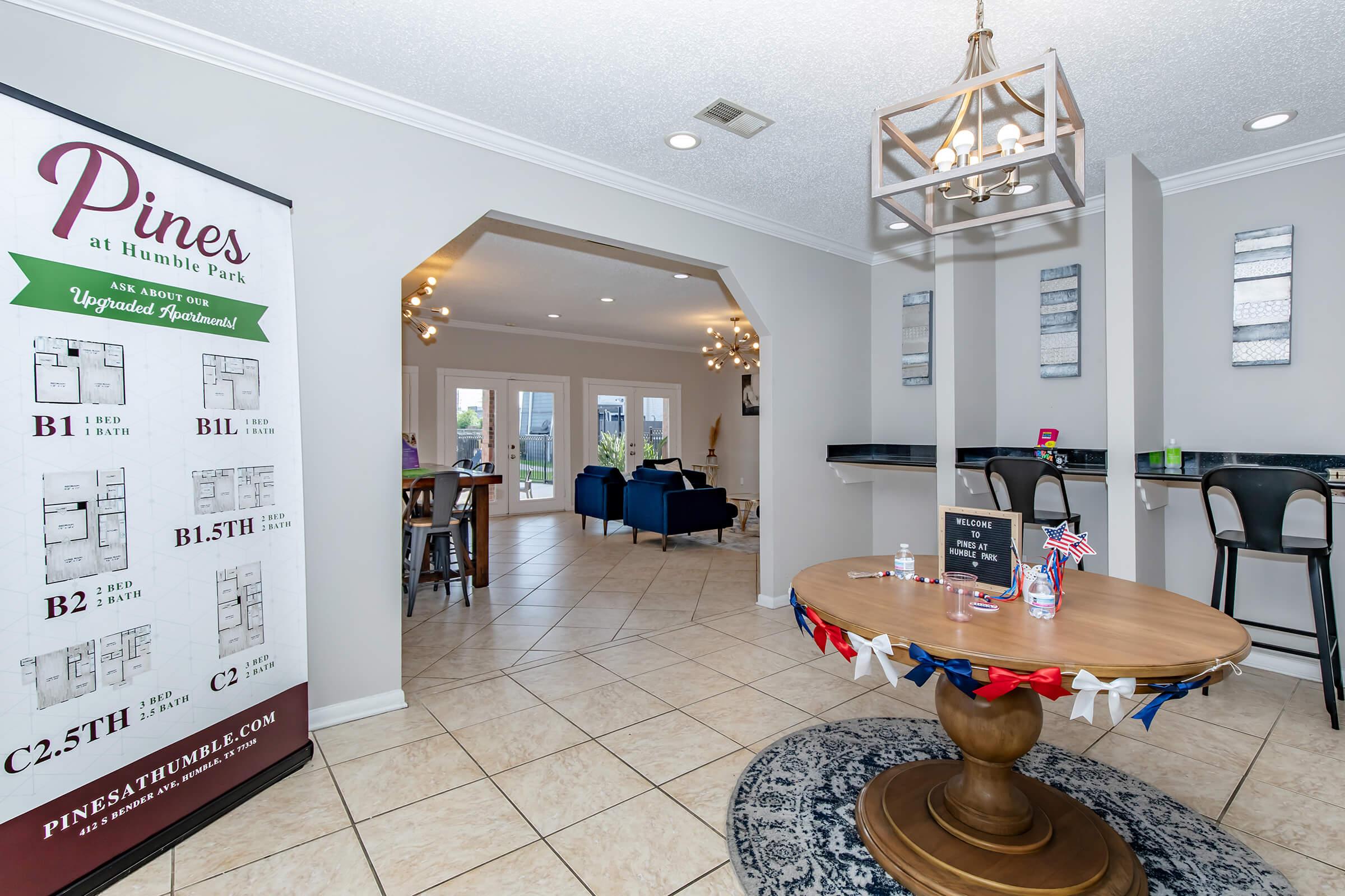A well-lit apartment leasing office with a decorative round table featuring a welcome sign and patriotic decor. A large poster on the wall displays floor plans for various apartment layouts. The space leads to a cozy lounge area with blue couches, large windows, and modern light fixtures.