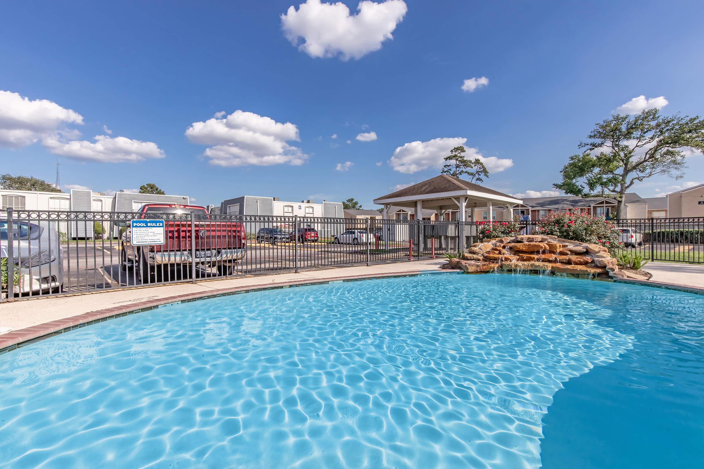 A clear blue swimming pool with a rock waterfall, surrounded by a black metal fence. In the background, there are several trailers and parked vehicles, along with a covered gazebo area. The sky is bright with fluffy white clouds.