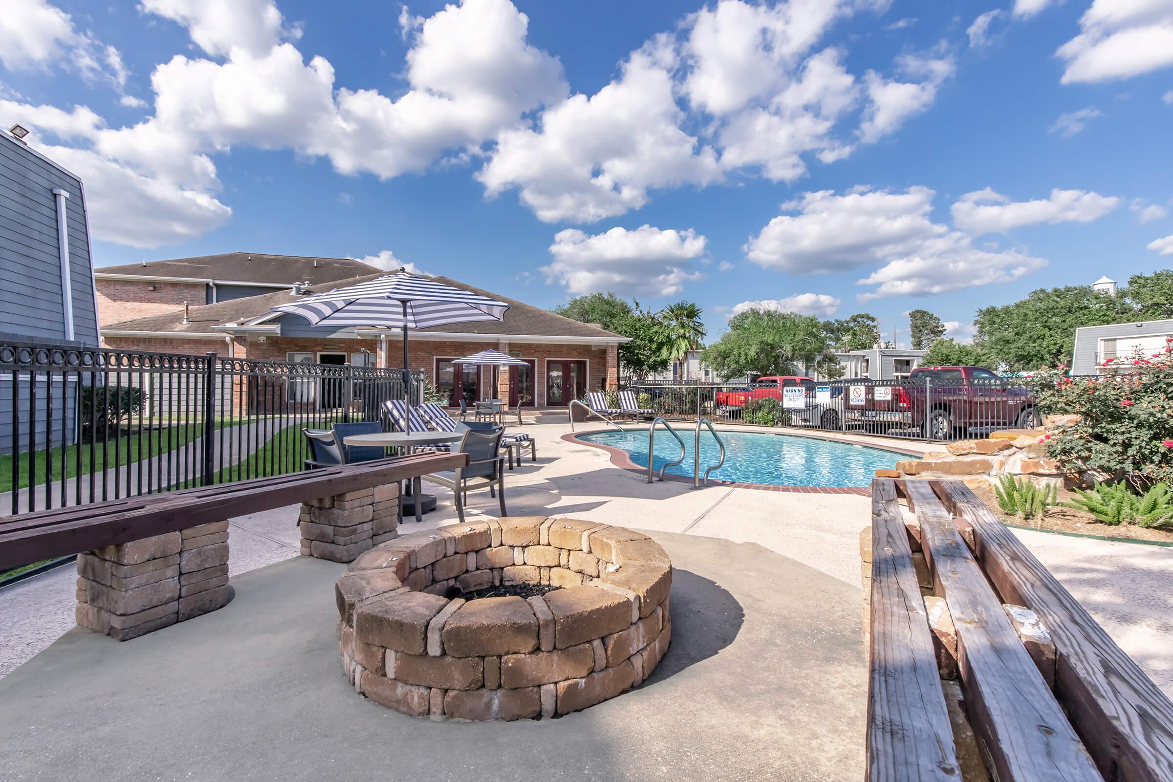 A serene outdoor pool area featuring a sparkling blue pool surrounded by lounge chairs, umbrellas, and a stone fire pit. Lush landscaping and a clear sky with fluffy clouds create a relaxing atmosphere. A building with multiple doors is visible in the background, enhancing the inviting ambiance.