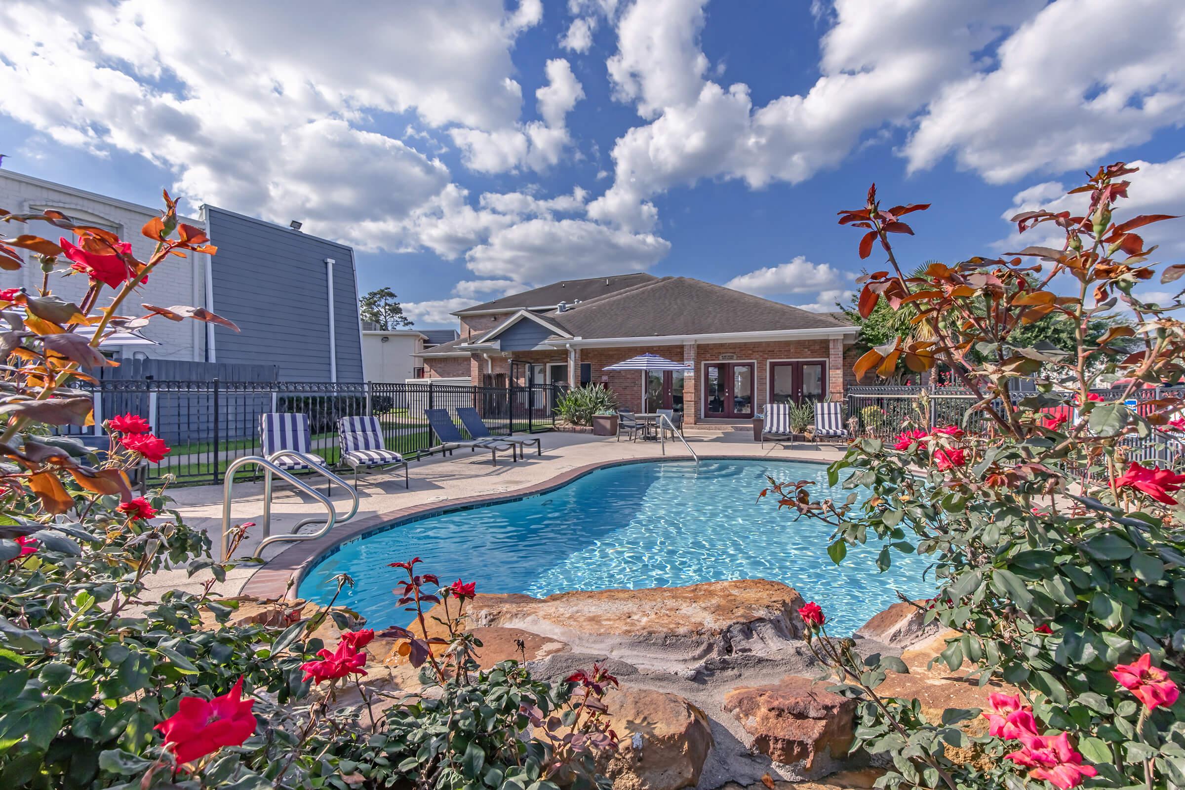 A serene outdoor swimming pool surrounded by colorful flowers and lush greenery. Sun loungers are positioned around the pool, and a patio area is visible in the background with a building, all under a bright blue sky with fluffy white clouds.