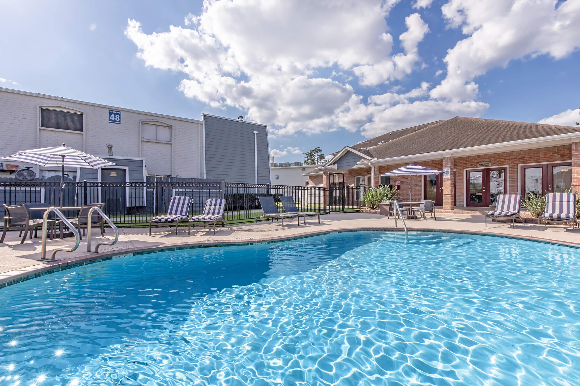 A sparkling blue swimming pool surrounded by lounge chairs and umbrellas, set against a backdrop of residential buildings and a partly cloudy sky. The area is well-maintained, inviting relaxation and enjoyment by the water.